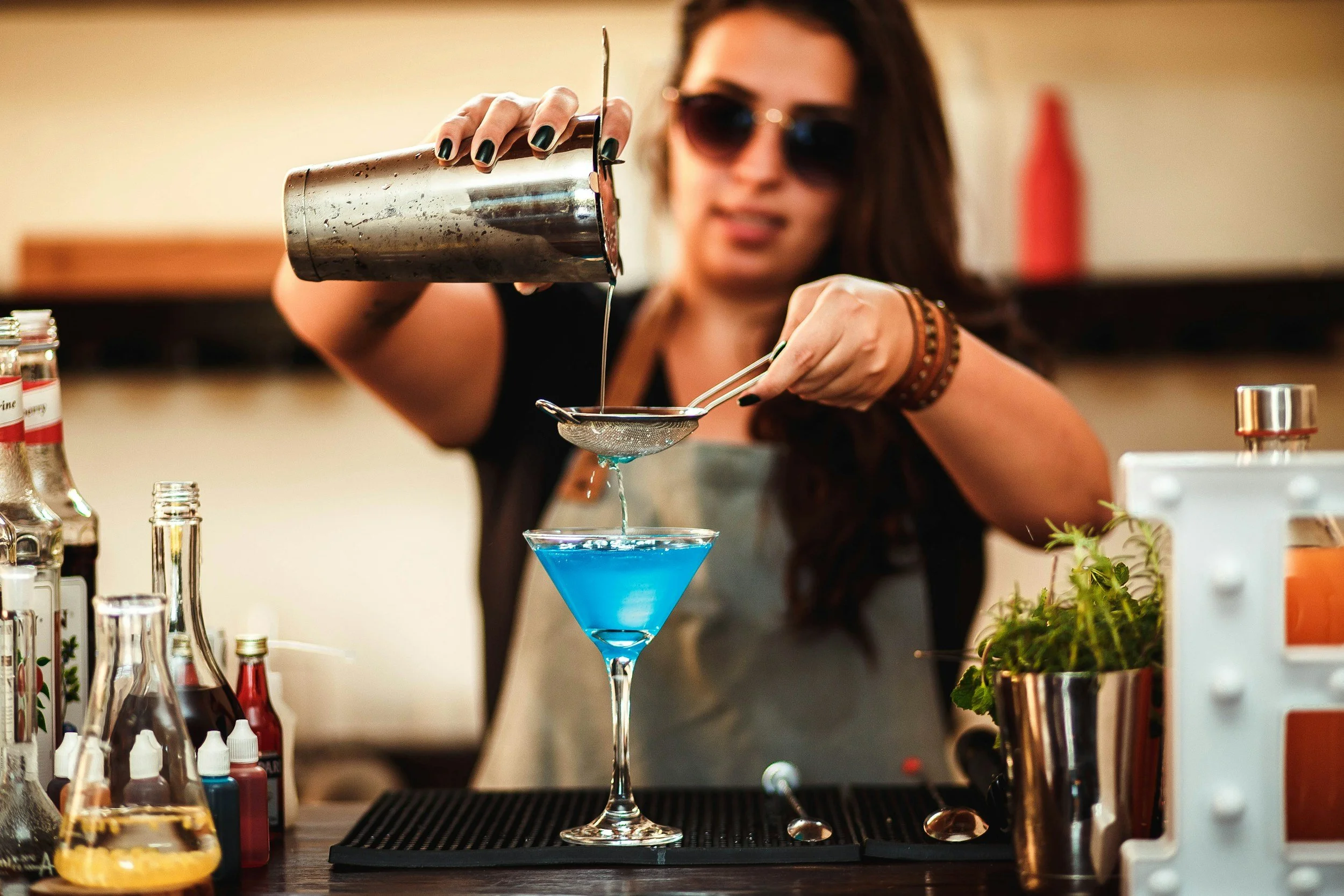 A woman with long dark hair and sunglasses pouring a blue cocktail through a strainer into a martini glass in a bar setting with various bottles and bar tools around her.
