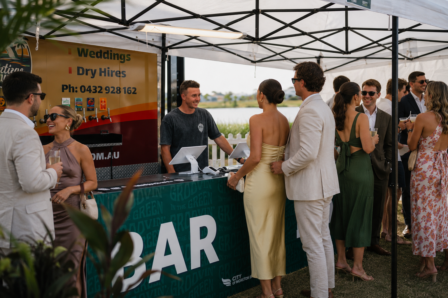 People gathered at a bar booth at an outdoor event, dressed in formal attire, with a water and landscape background.