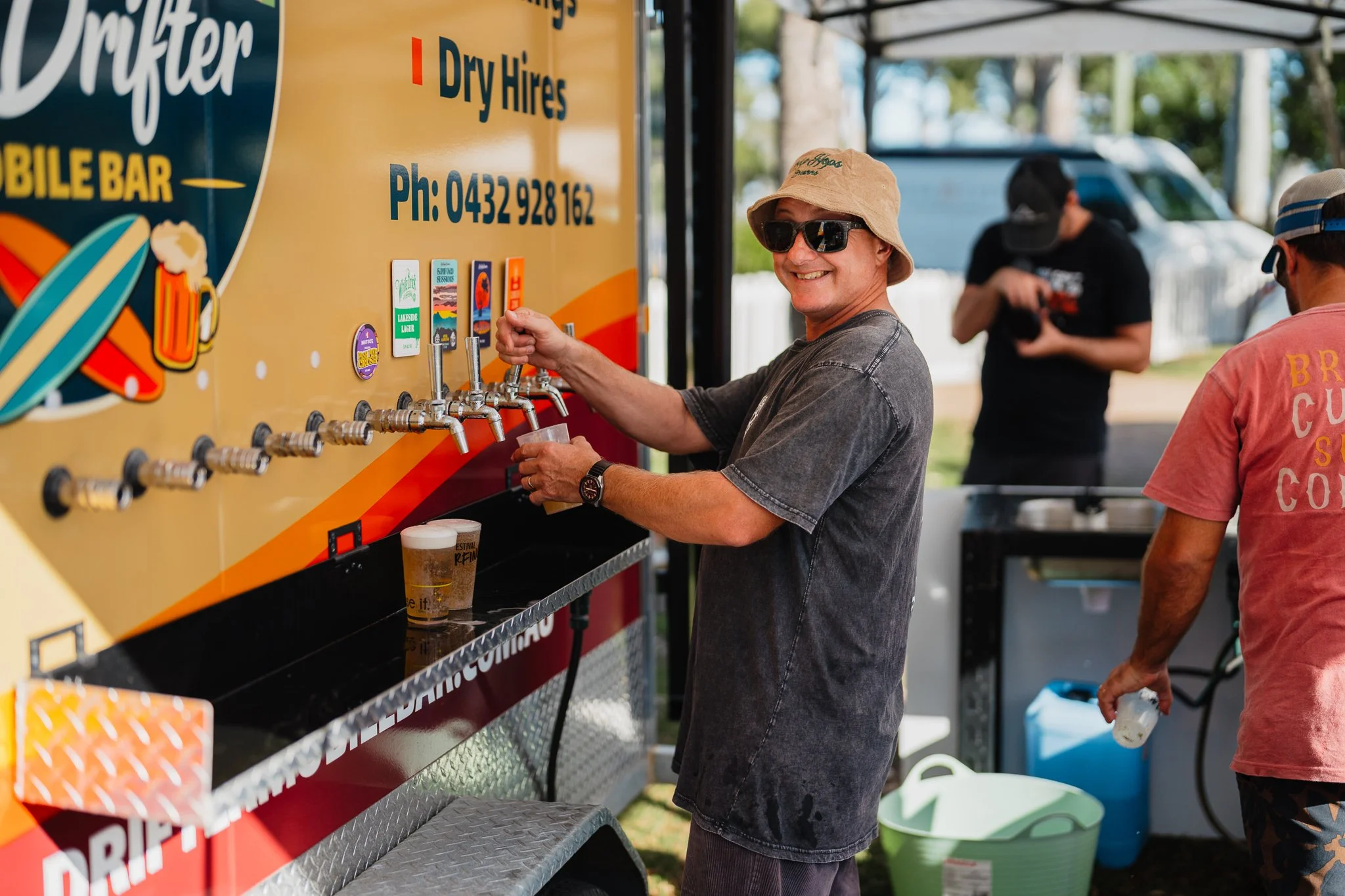 A man wearing sunglasses and a beige hat pours a beer from a tap at a mobile bar. He is smiling and standing outdoors with trees in the background. Another man in a pink t-shirt and a cap is visible nearby.