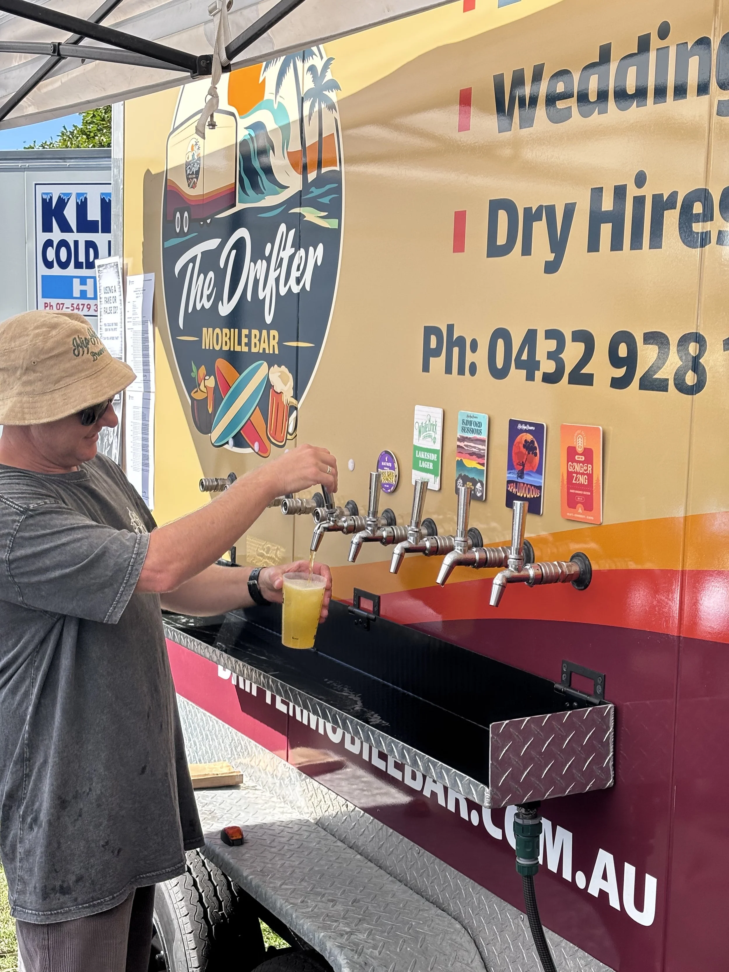 Person pouring a drink from taps on a mobile bar truck labeled 'The Drifter' with an illustration of a van, palm trees, and surfboards. The truck advertises wedding and dry hire services, with a phone number and a row of colorful beer coaster signs.
