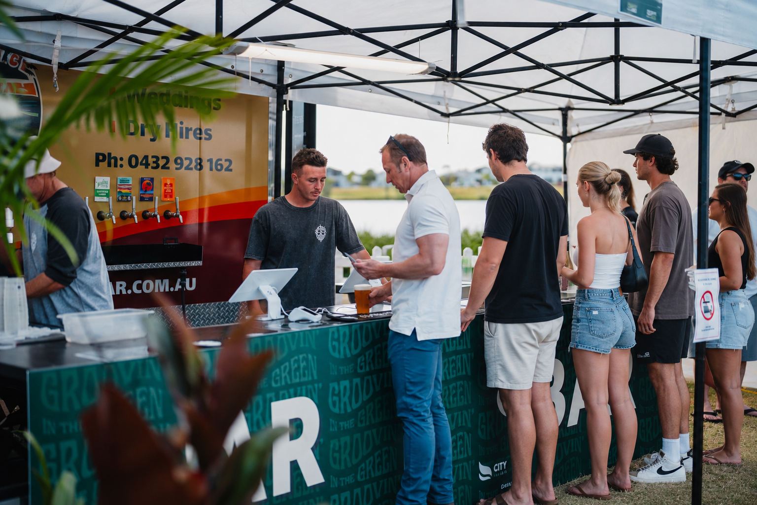People are standing in line at an outdoor food stall under a canopy, ordering drinks with a water body and cloudy sky in the background.