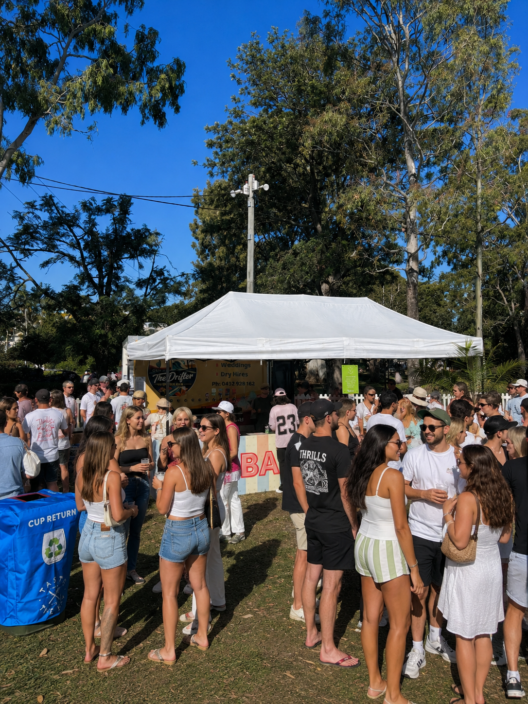 A crowd of people gathered outdoors under a white tent, with trees and a clear blue sky in the background, at what appears to be a festival or outdoor event.
