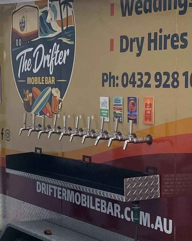 A mobile bar trailer called 'The Drifter' with multiple beer taps mounted on a wall, and a black serving area underneath. The trailer features colorful graphics including surfboards, waves, and a beer mug, with a large sign providing contact information and services for weddings and dry hire.