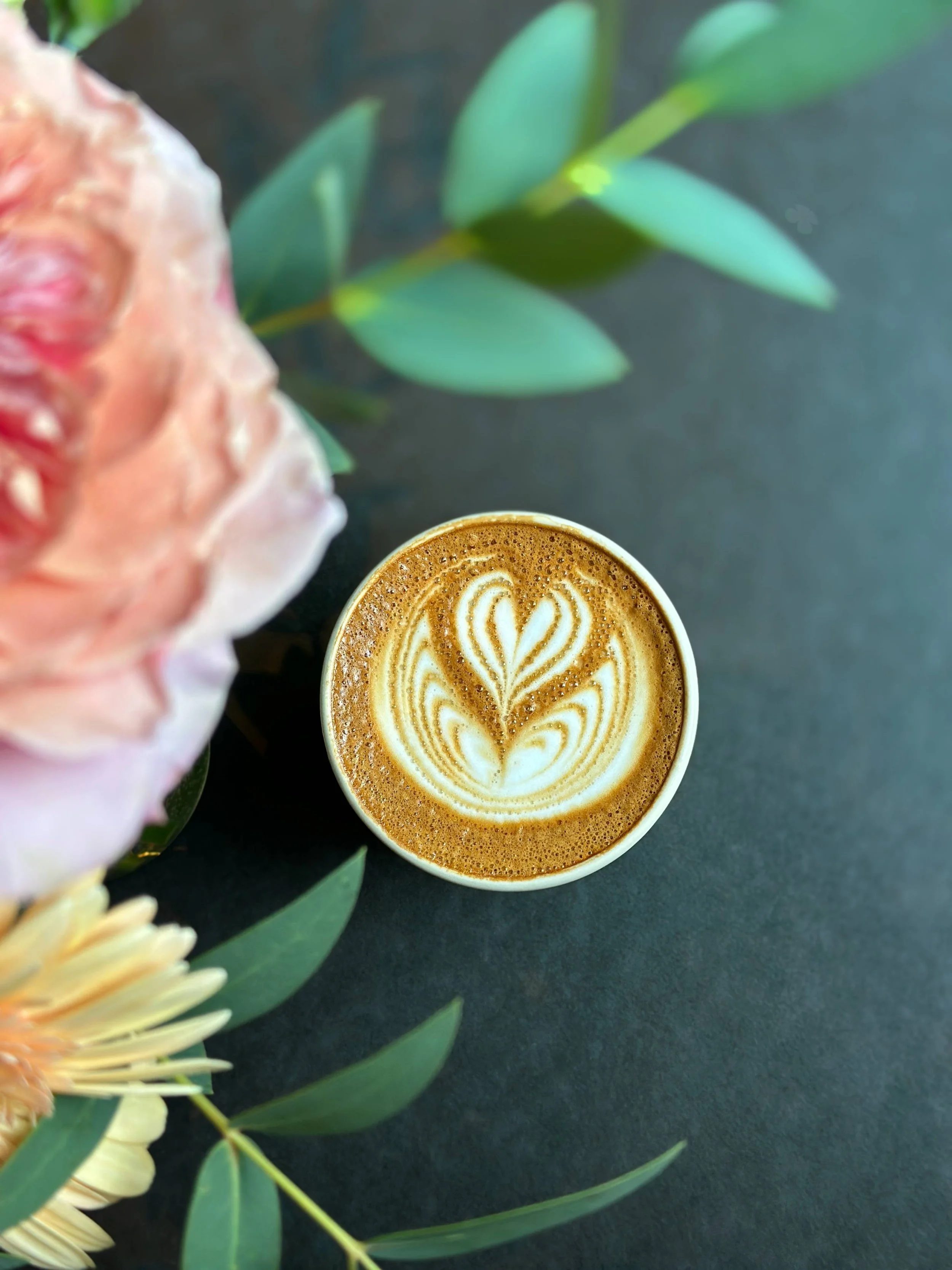 A top-down view of a cup of latte with foam art in the shape of a heart and floral pattern, surrounded by pink and white flowers and green leaves on a dark surface.