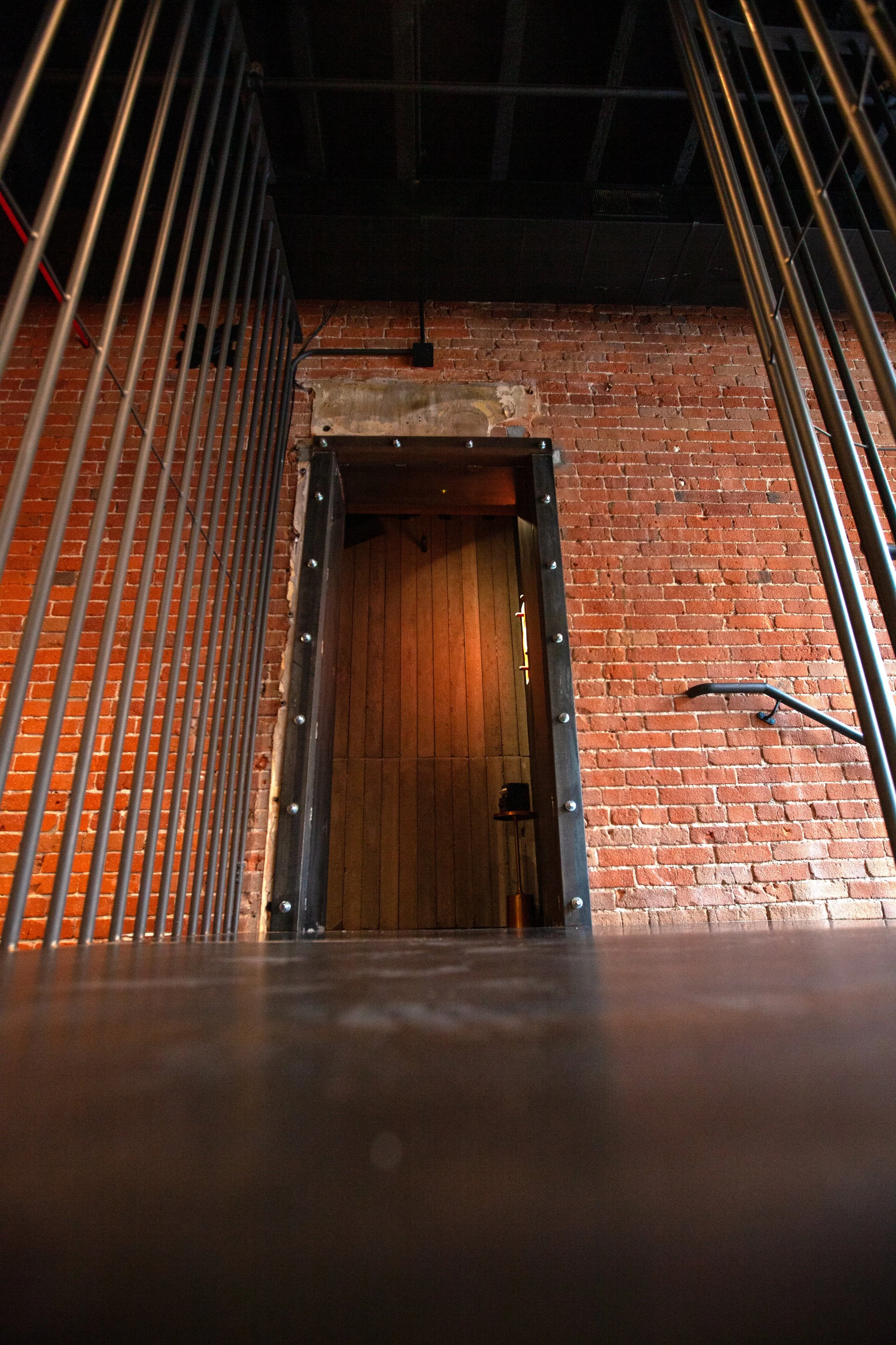 View of an industrial-style brick wall with a doorway framed with metal bolts, leading into a room with wooden walls and a small table with a dark object.
