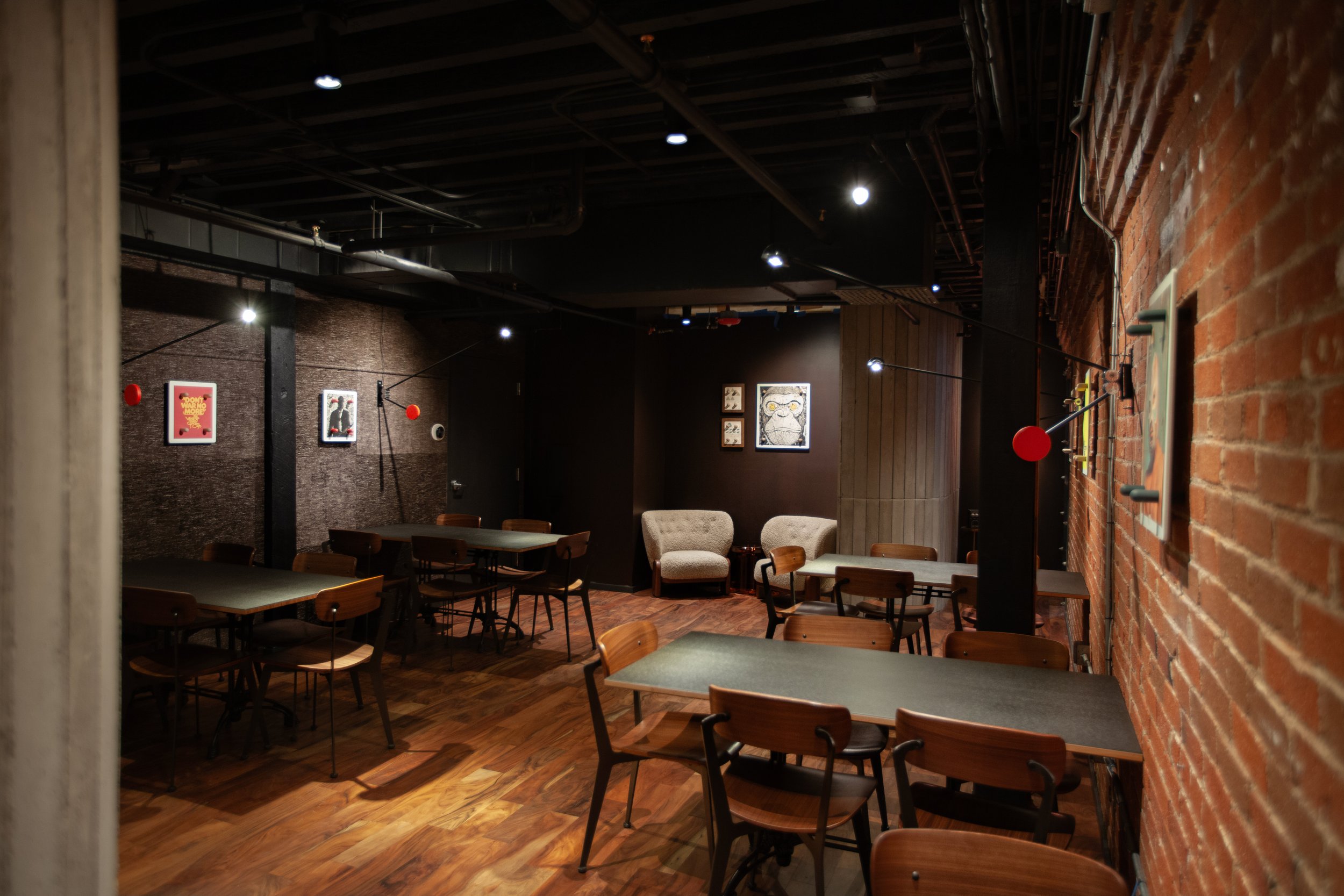Interior of a dimly lit cafe or restaurant with wooden floors, brick and black walls, and chairs arranged around tables, some artwork hanging on the walls.