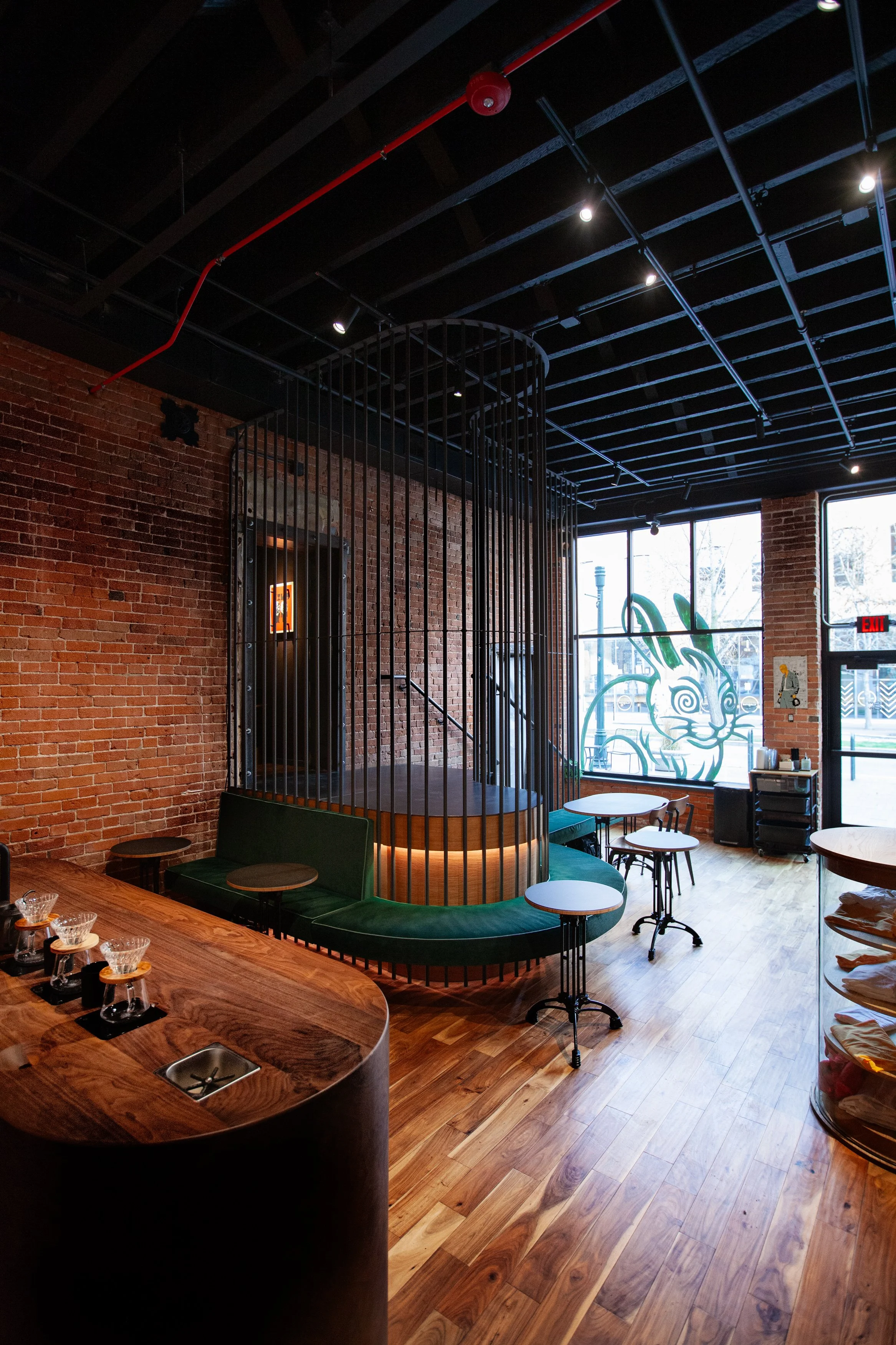 Interior of a cafe with brick walls, wooden flooring, green seating, and tables near large front windows with a dragon artwork.