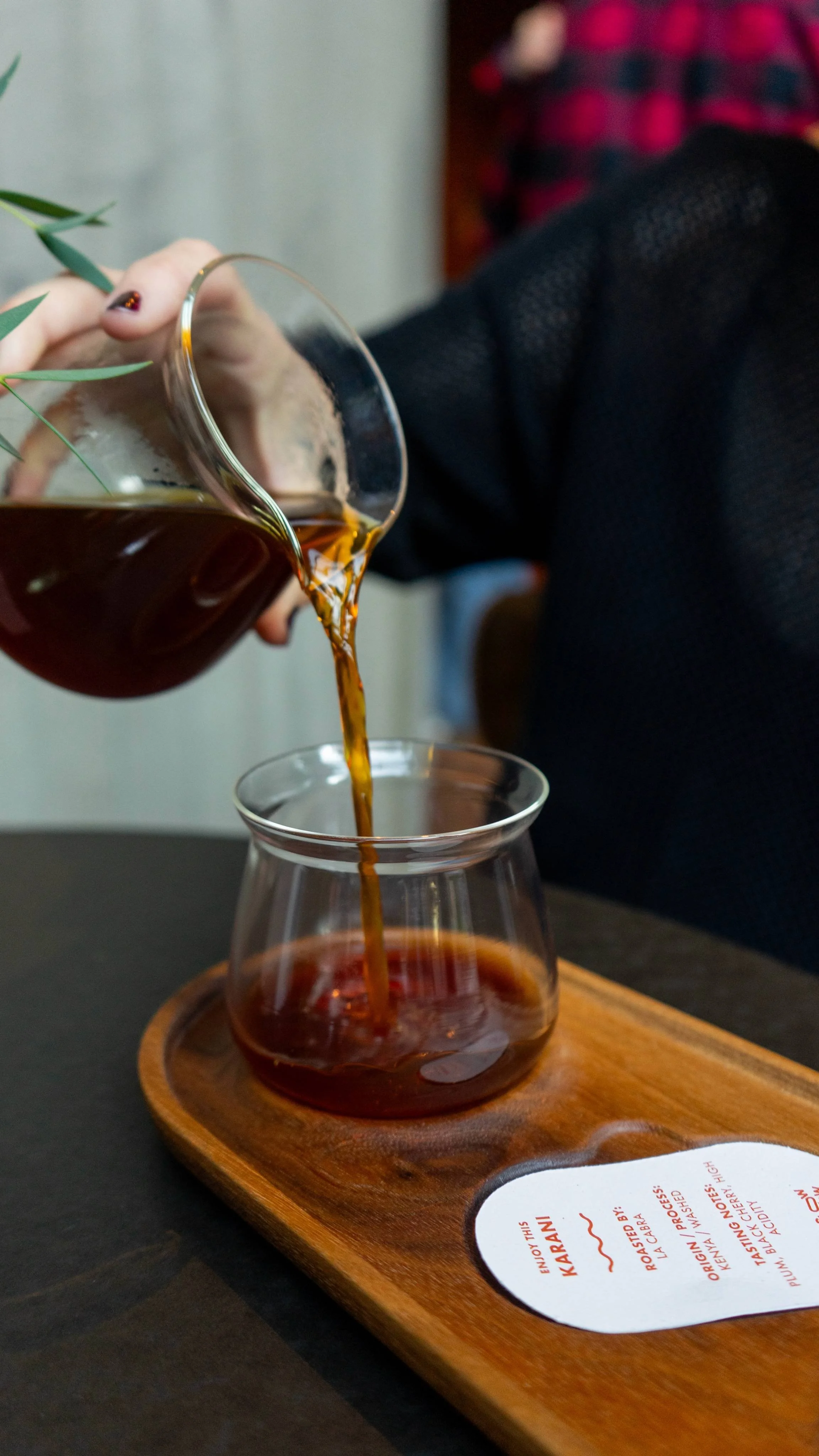 A person pouring rich specialty roasted pourover coffee into a glass cup placed on a wooden tray.