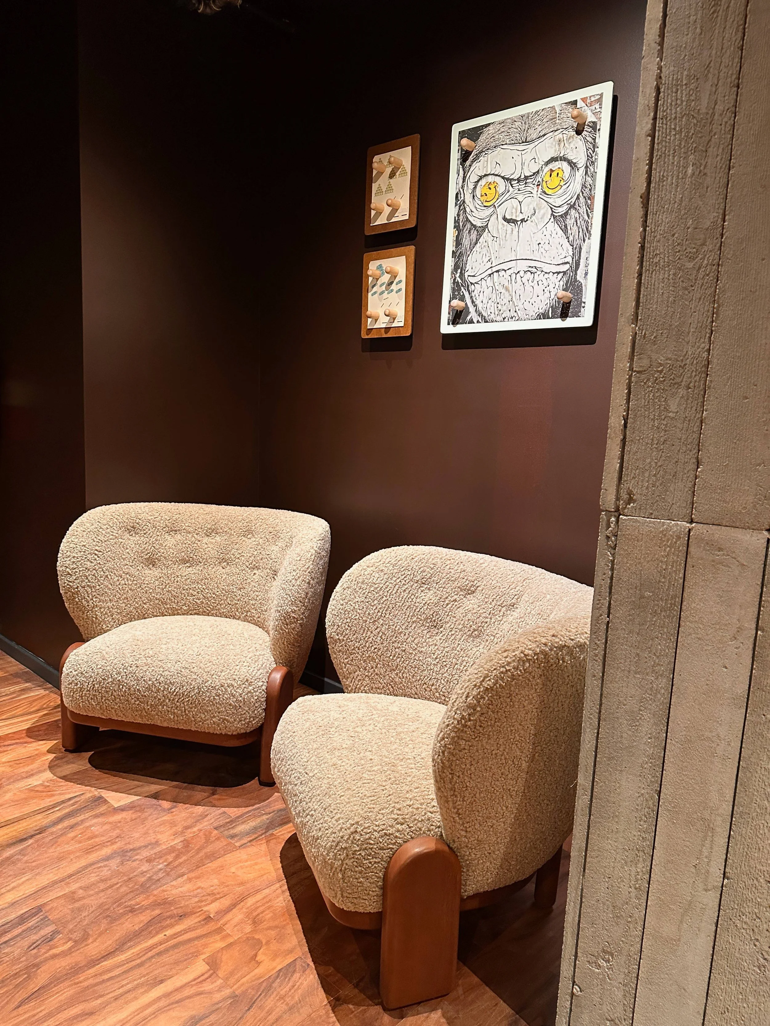 Two beige, textured, plush armchairs with wooden legs sit on a polished wooden floor against dark brown walls, with colorful artwork hanging above them.