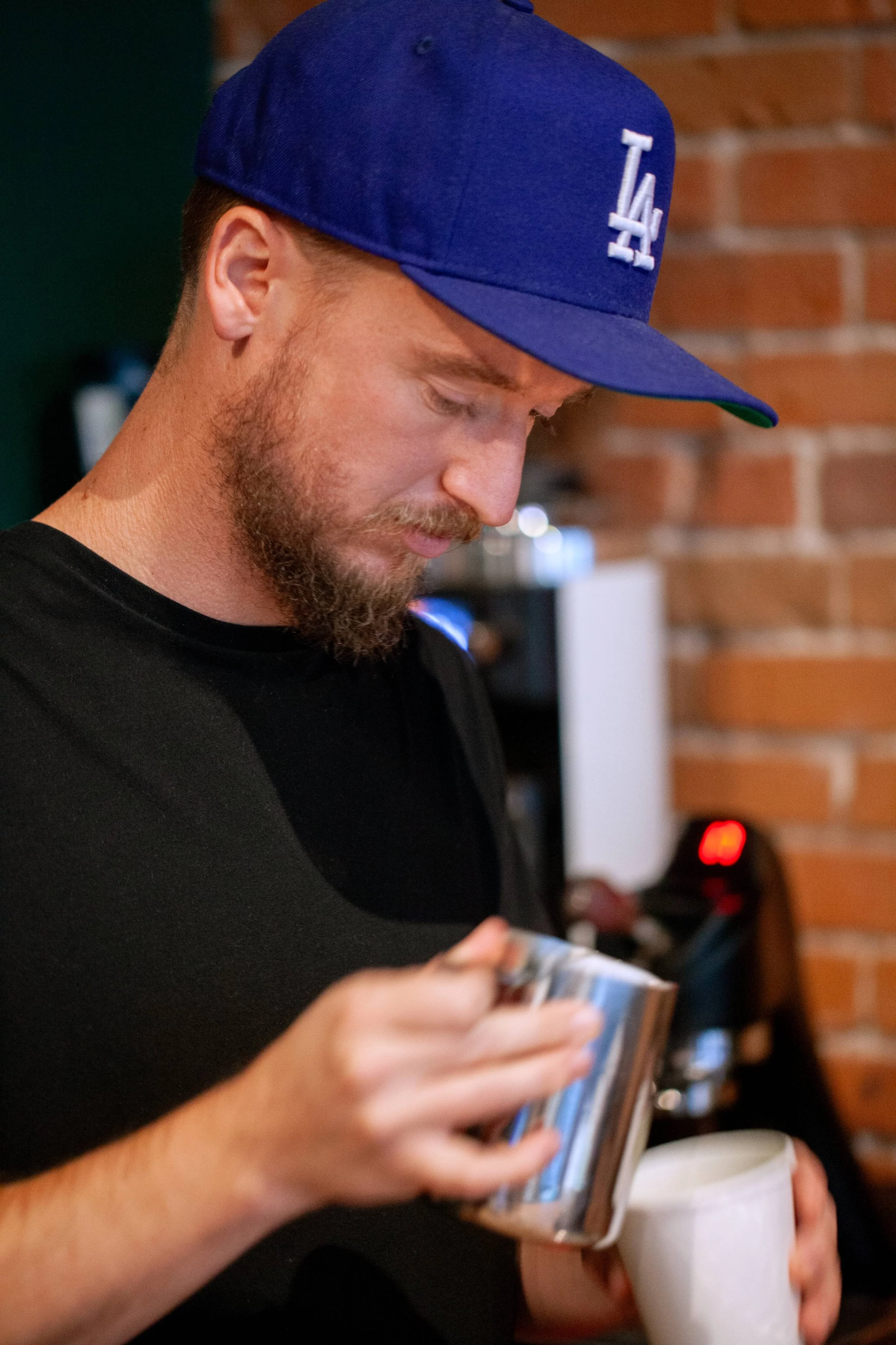 A man with a beard wearing a blue Los Angeles Dodgers cap, holding a white mug in one hand a milk carafe in the other, pouring a latte in front of a brick wall.