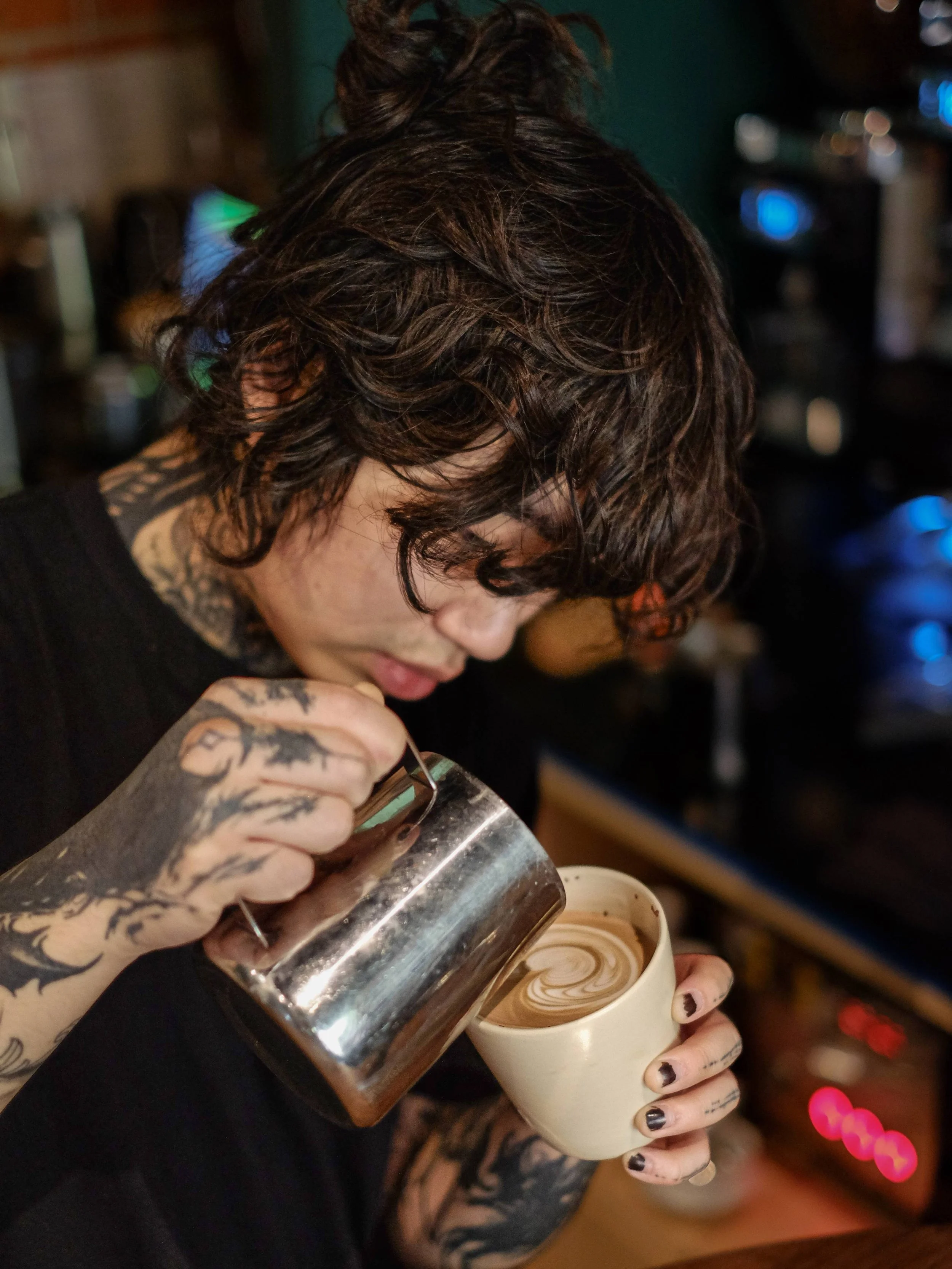 A person with tattoos and dark hair styles in a messy bob, pouring steamed milk into a coffee cup to create latte art.