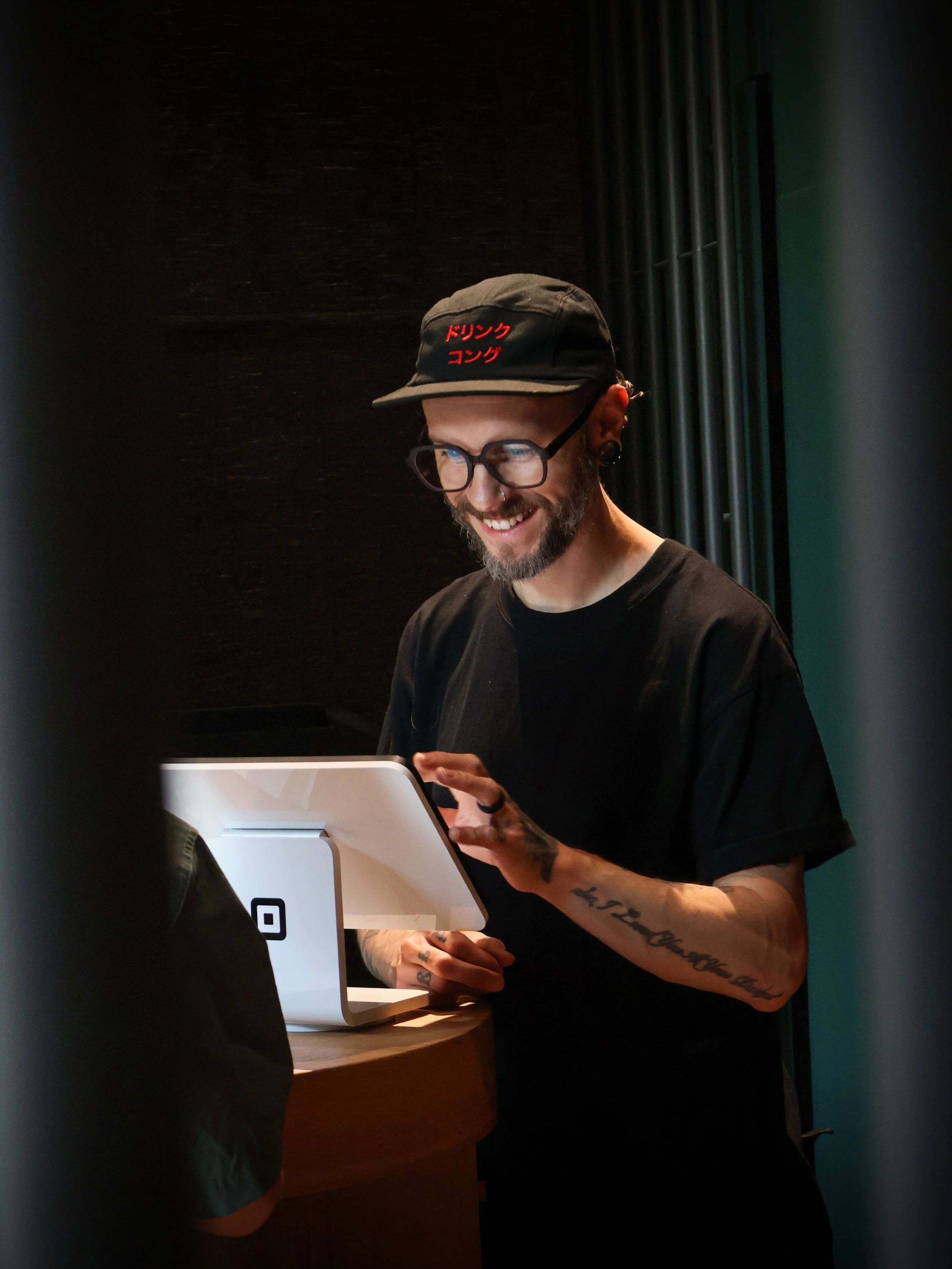 A man with tattoos, glasses, a beard, and piercings, wearing a black cap with red Japanese characters, smiling while using a white tablet or register at a counter in a dark room.