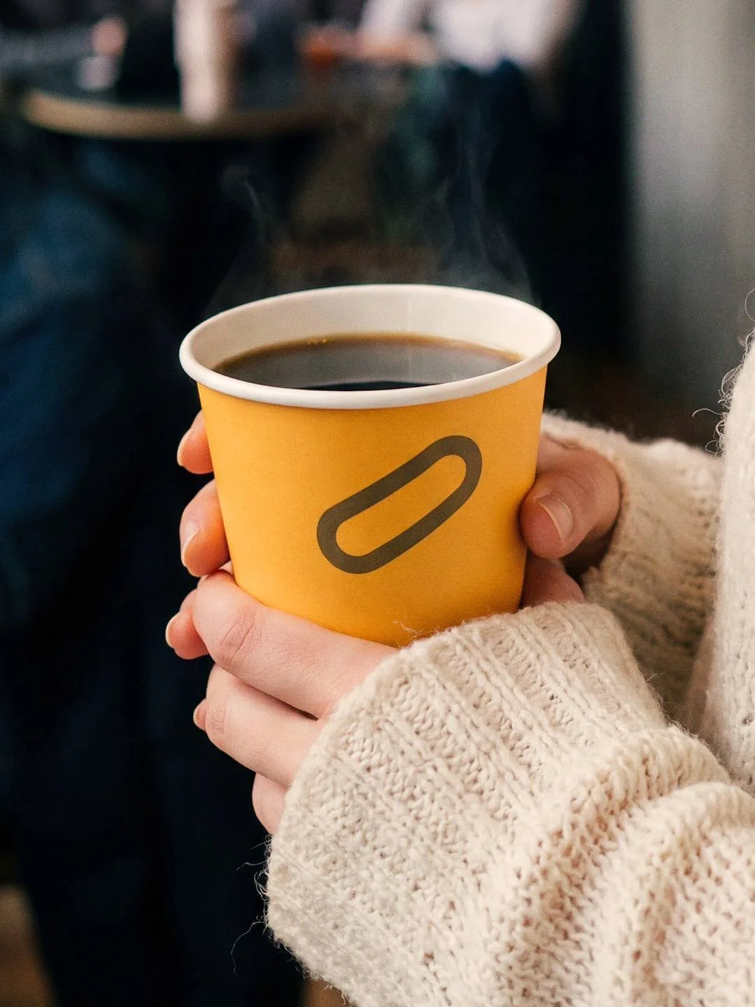 Person holding a steaming cup of black coffee in a yellow disposable cup with a printed logo, wearing a beige knitted sweater.