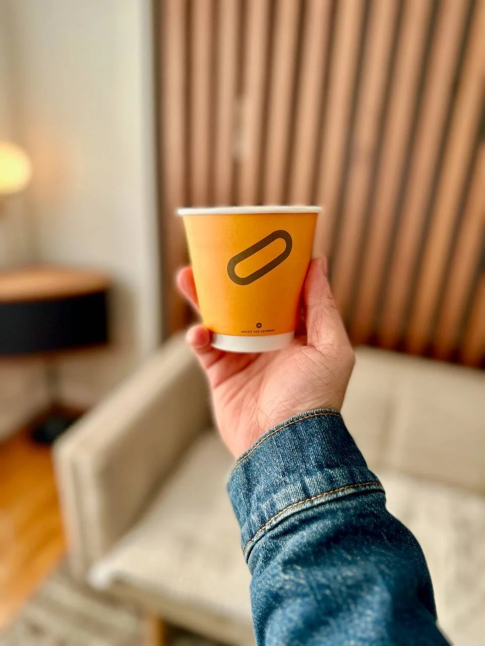 Hand holding a yellow paper cup with a minimalist Slow by Slow logo, in a room with wooden paneling and a beige couch in the background.