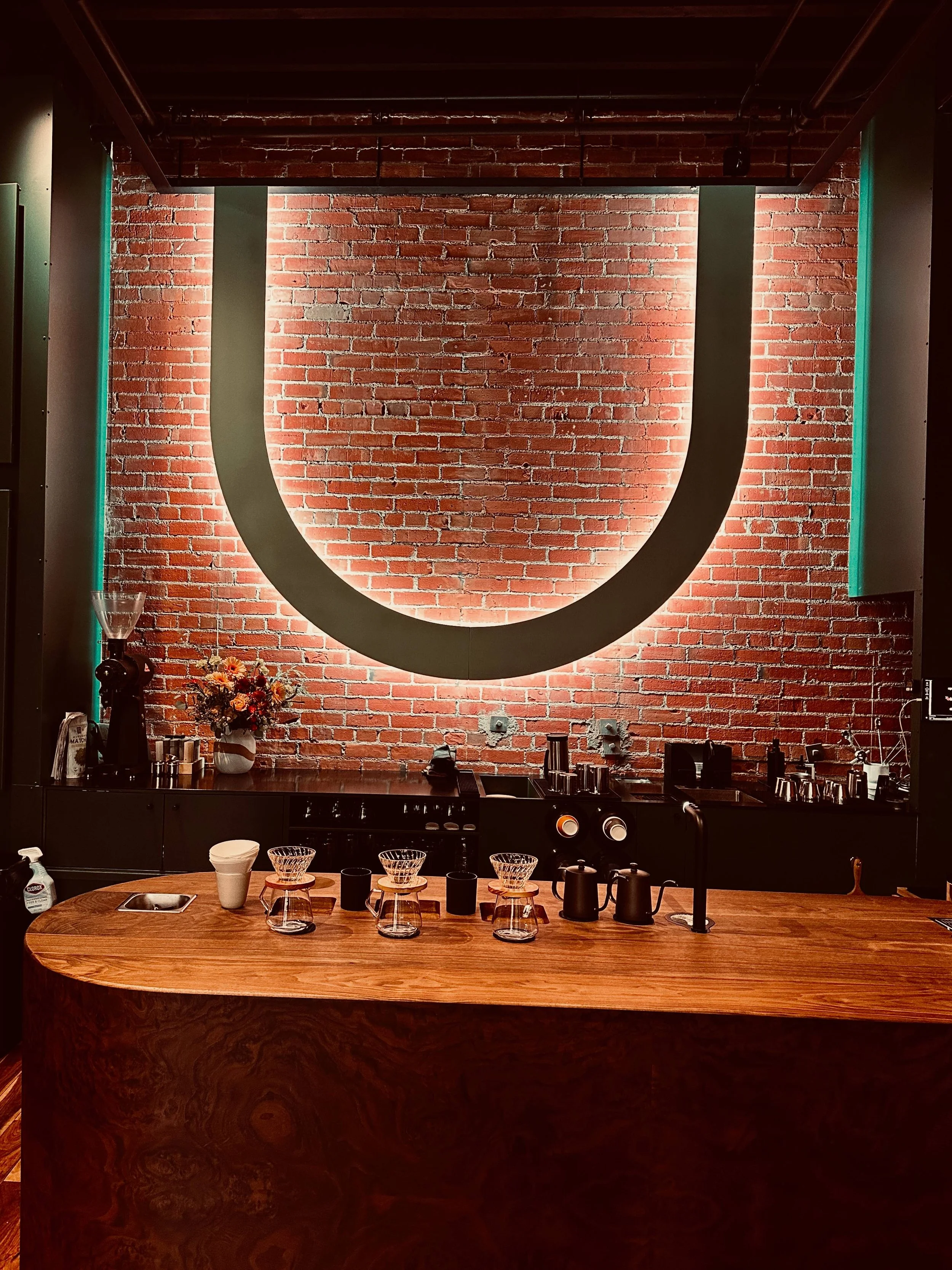 A coffee bar with a curved wooden counter, four coffee glasses, and black coffee cups, set against a brick wall with a large illuminated 'U' shape in the background.