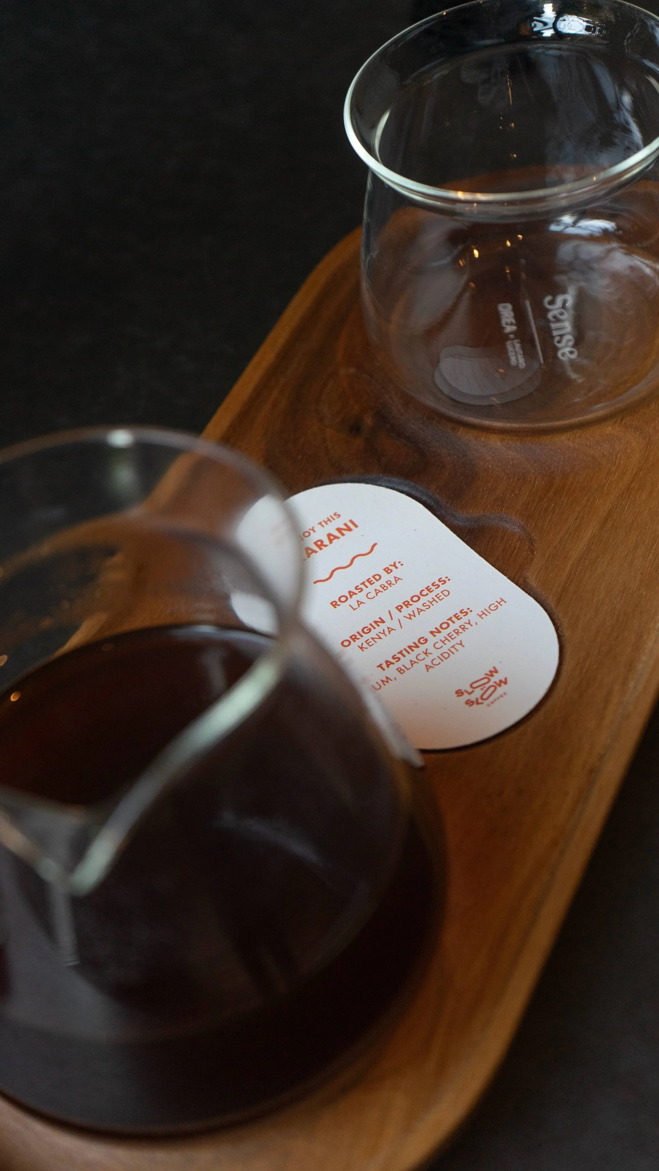 Two glass cups filled with dark coffee resting on a wooden tray with a label describing the coffee origin, process, tasting notes, and roasting details.