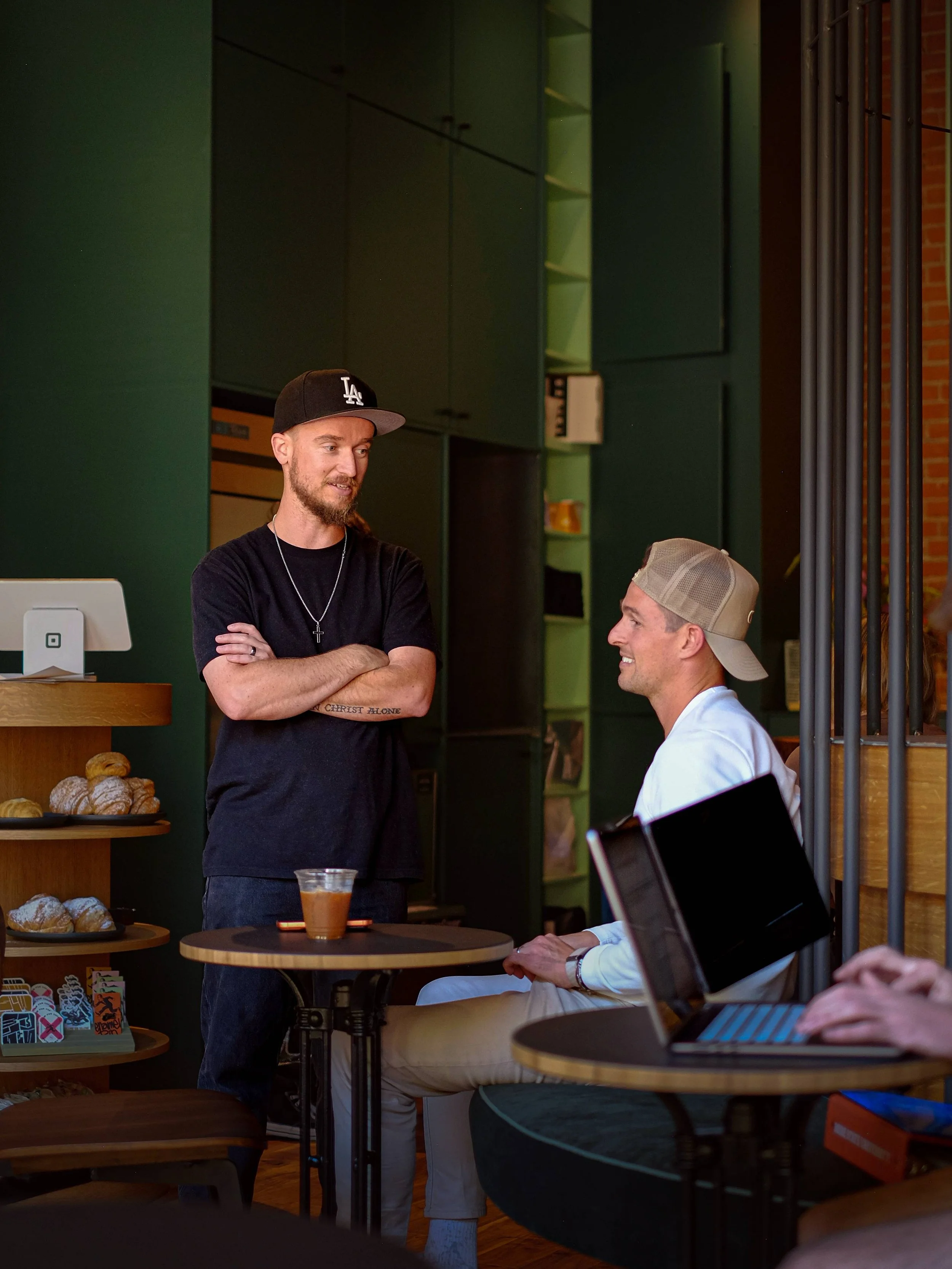 Two men conversing in a cafe; one standing with crossed arms and the other sitting; both smiling, with a laptop and coffee on the tables.