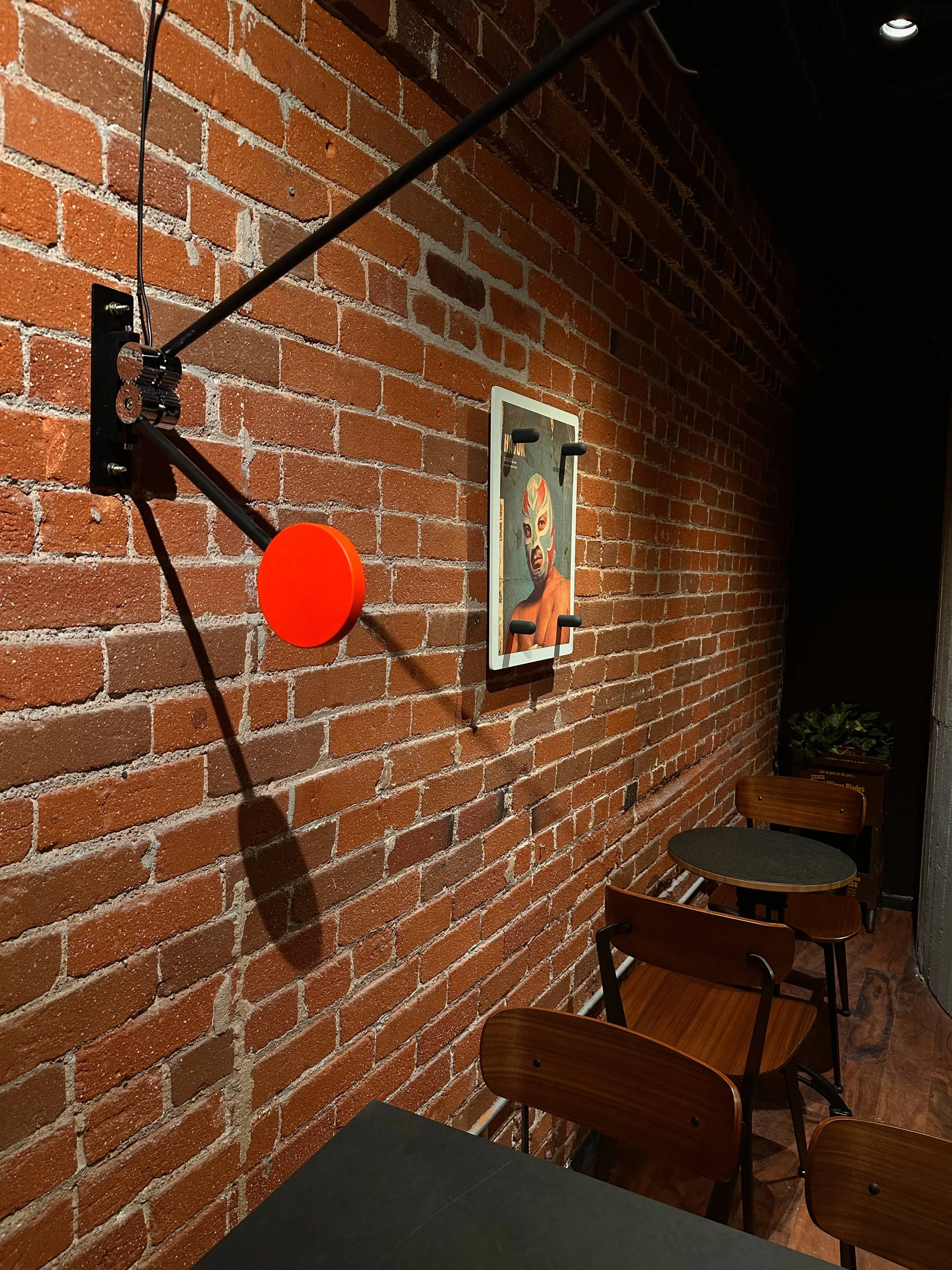 A brick wall with a black structural element and a mounted orange disc, a small framed artwork of a man wearing a lucha libre mask, and a small round table with two wooden chairs nearby, along with a small plant in the corner.