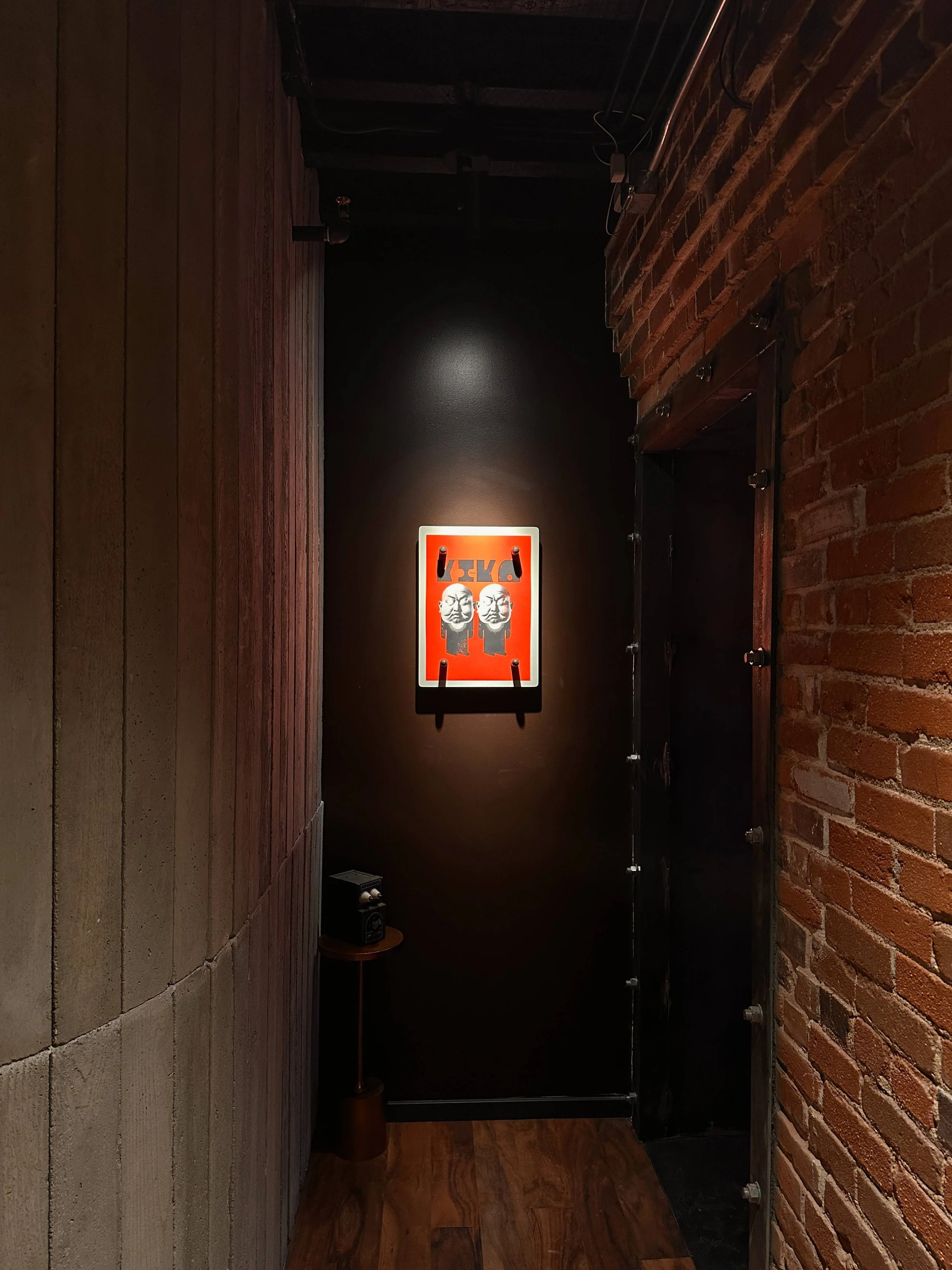A small hallway with dark walls and exposed brick, featuring a framed artwork of two faces and the word "KIKO" above in red, with a small speaker on a side table.