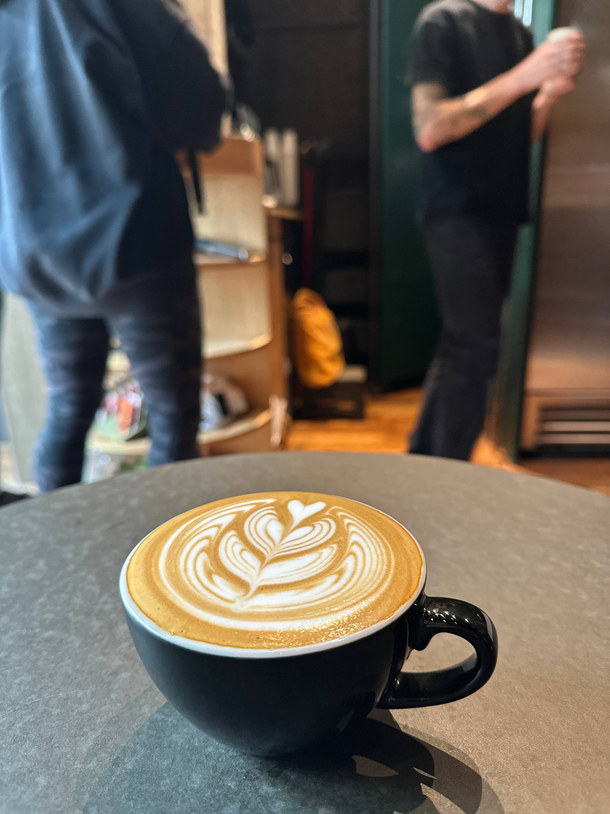 A cup of latte with intricate latte art on top, placed on a dark gray table in a cafe. In the background, two people are standing and conversing, one with tattoos and a black shirt, the other with a hoodie and jeans.