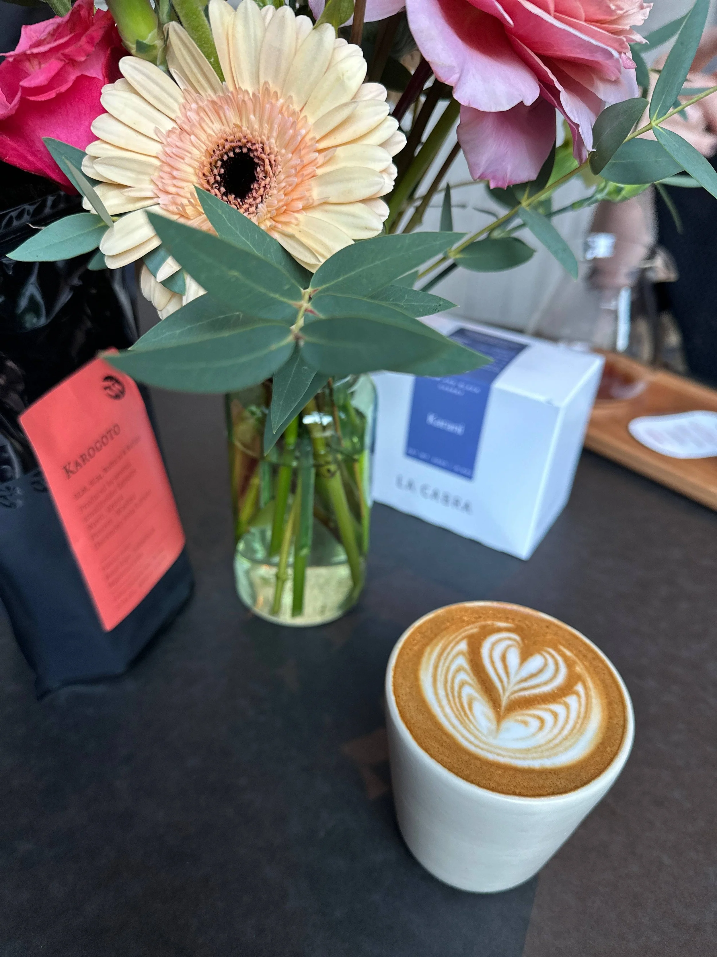 A cup of latte with heart-shaped latte art, a clear glass vase with pink and cream roses, peonies, and green leaves, a black bag with a red tag, and a box of tea on a dark surface.