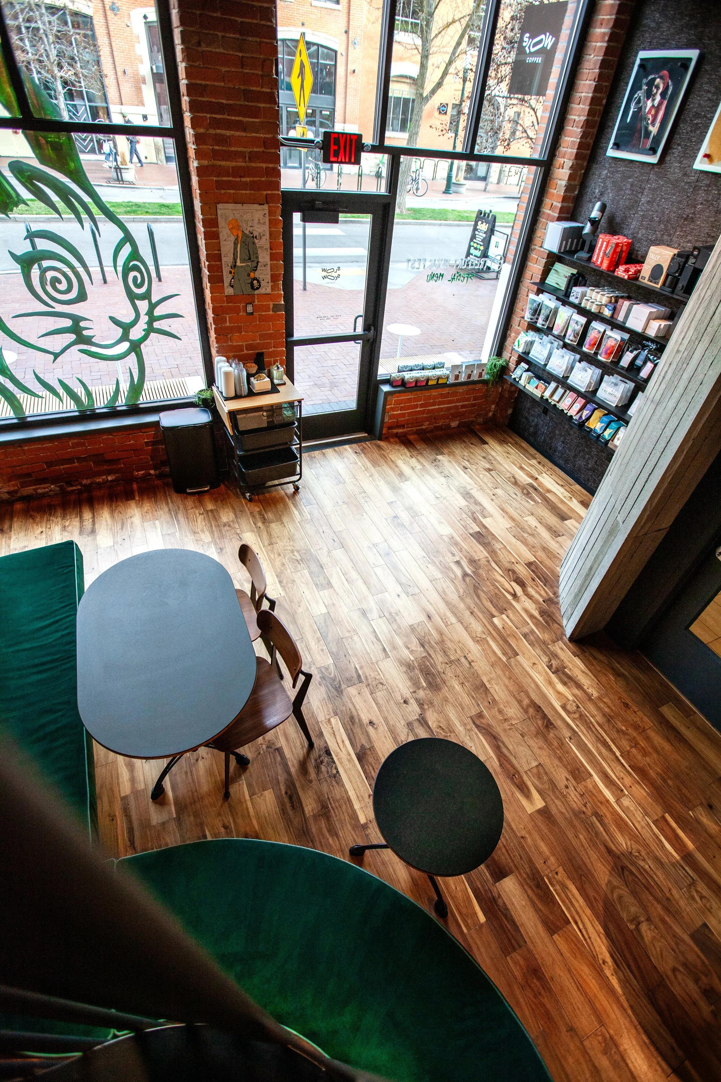 Interior of a coffee shop with wooden flooring, tall brick walls, large glass windows, a small black table with two wooden chairs, a small black round side table, shelves with books and products, and a large window decal of a hypnotized rabbit.