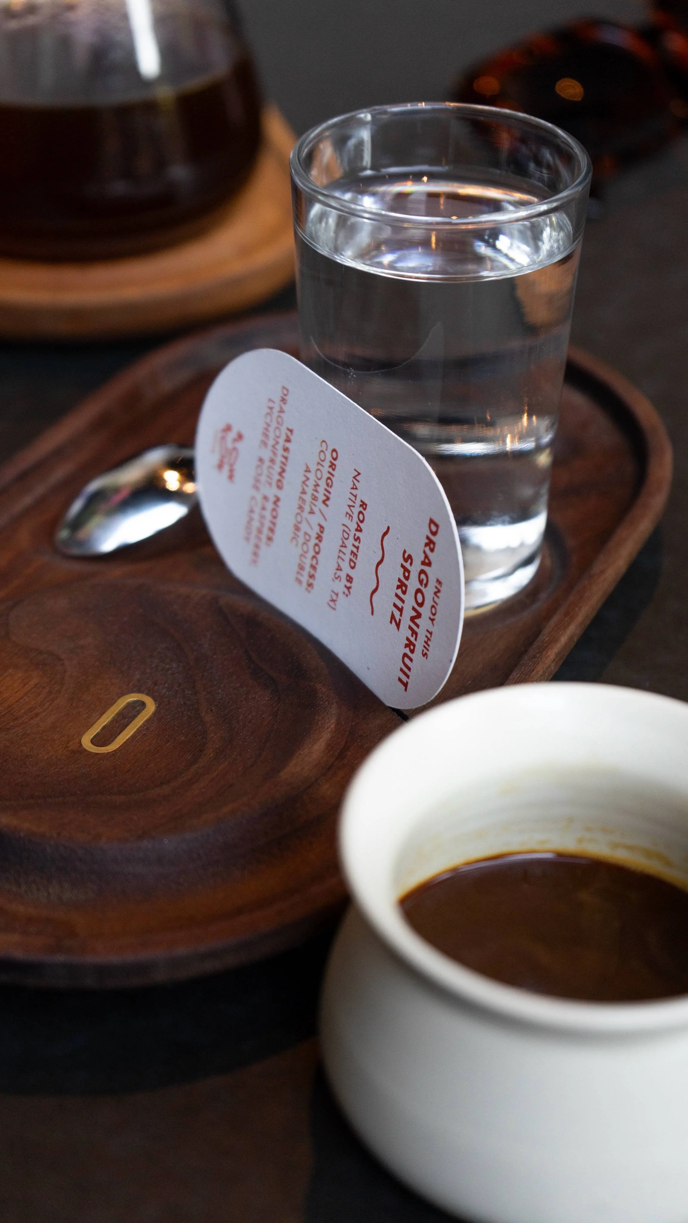 A glass of water, a spoon, a small white cup filled with espresso and a wooden tray with a paper tag that reads 'Dragonfly Spiritz' placed on top of the tray.