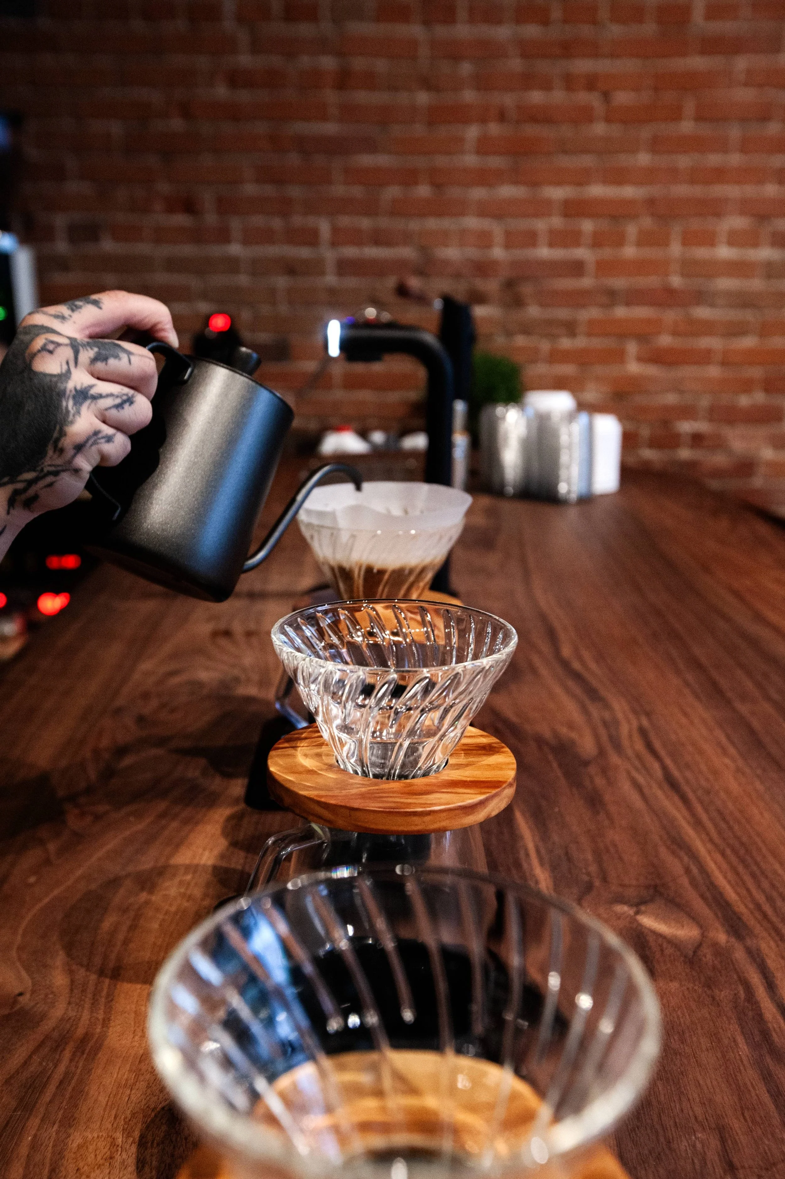 Barista's hand pouring coffee into glass dripper on wooden stand at a coffee bar with a brick wall background.