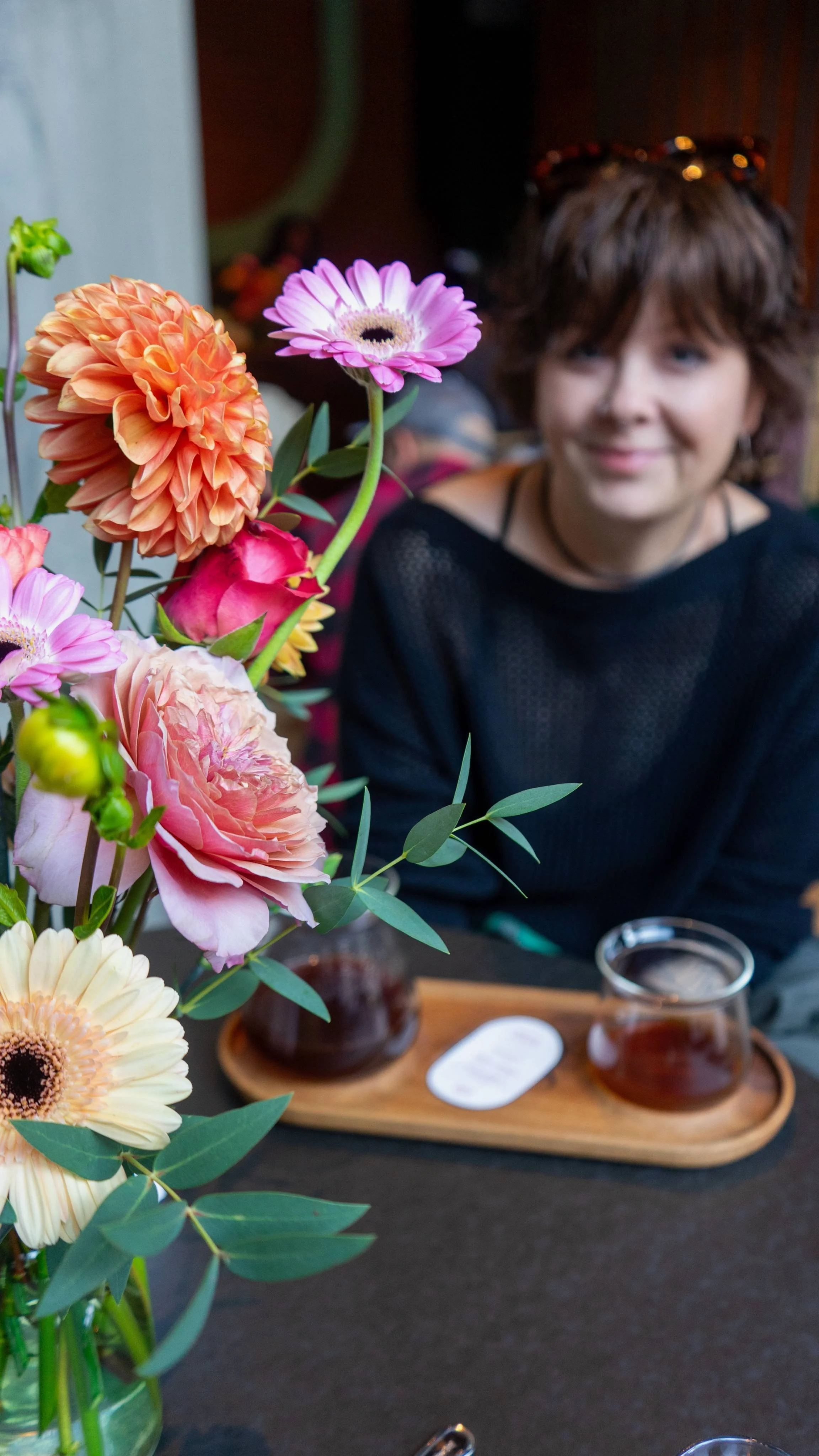 A woman with short, brown hair and sunglasses on her head sits at a table with a large bouquet of colorful flowers in the foreground. The flowers include pink, peach, and cream-colored blooms, and the woman is smiling softly.