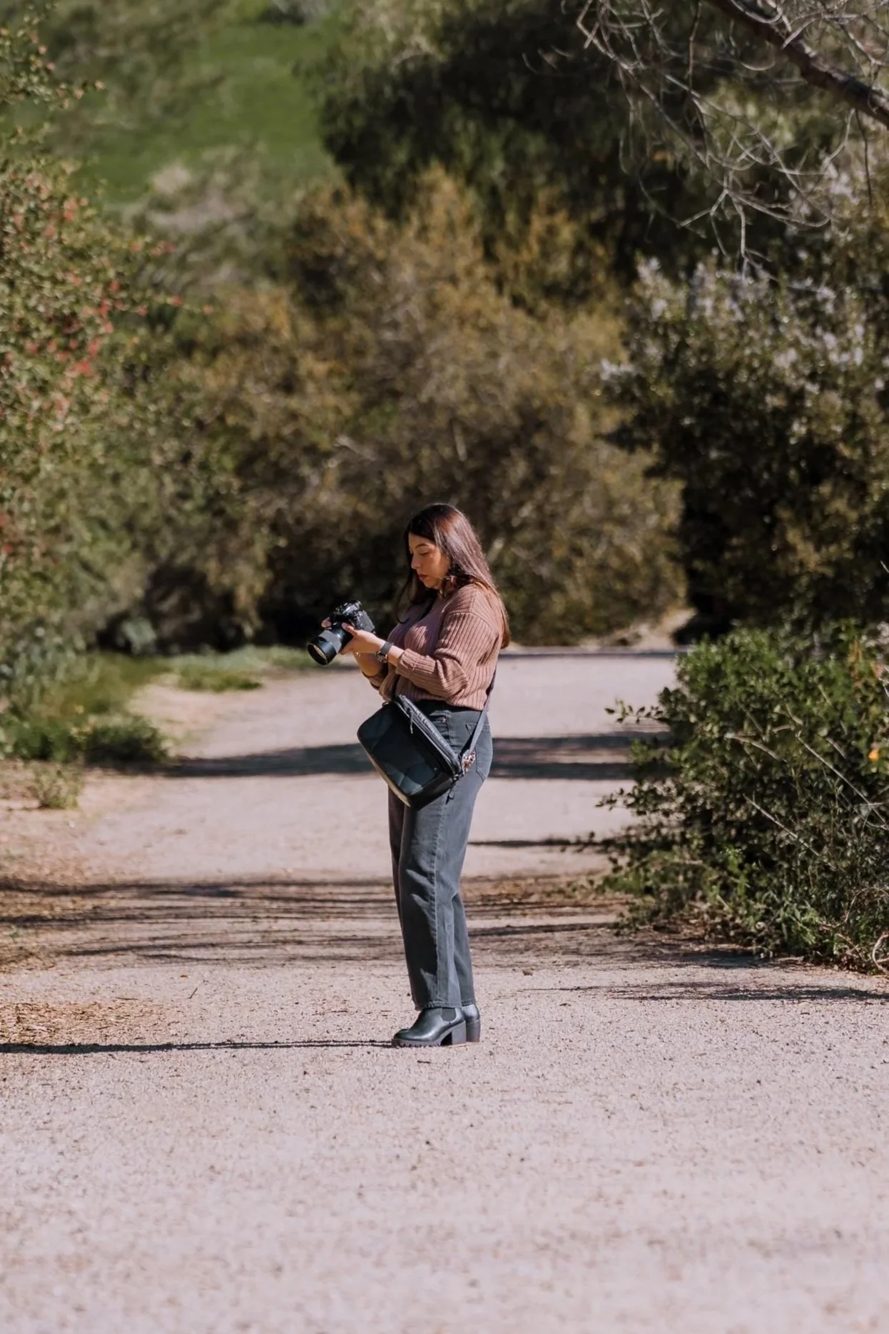 A woman standing on a dirt path in a park holding a camera and looking at it, surrounded by trees and greenery.