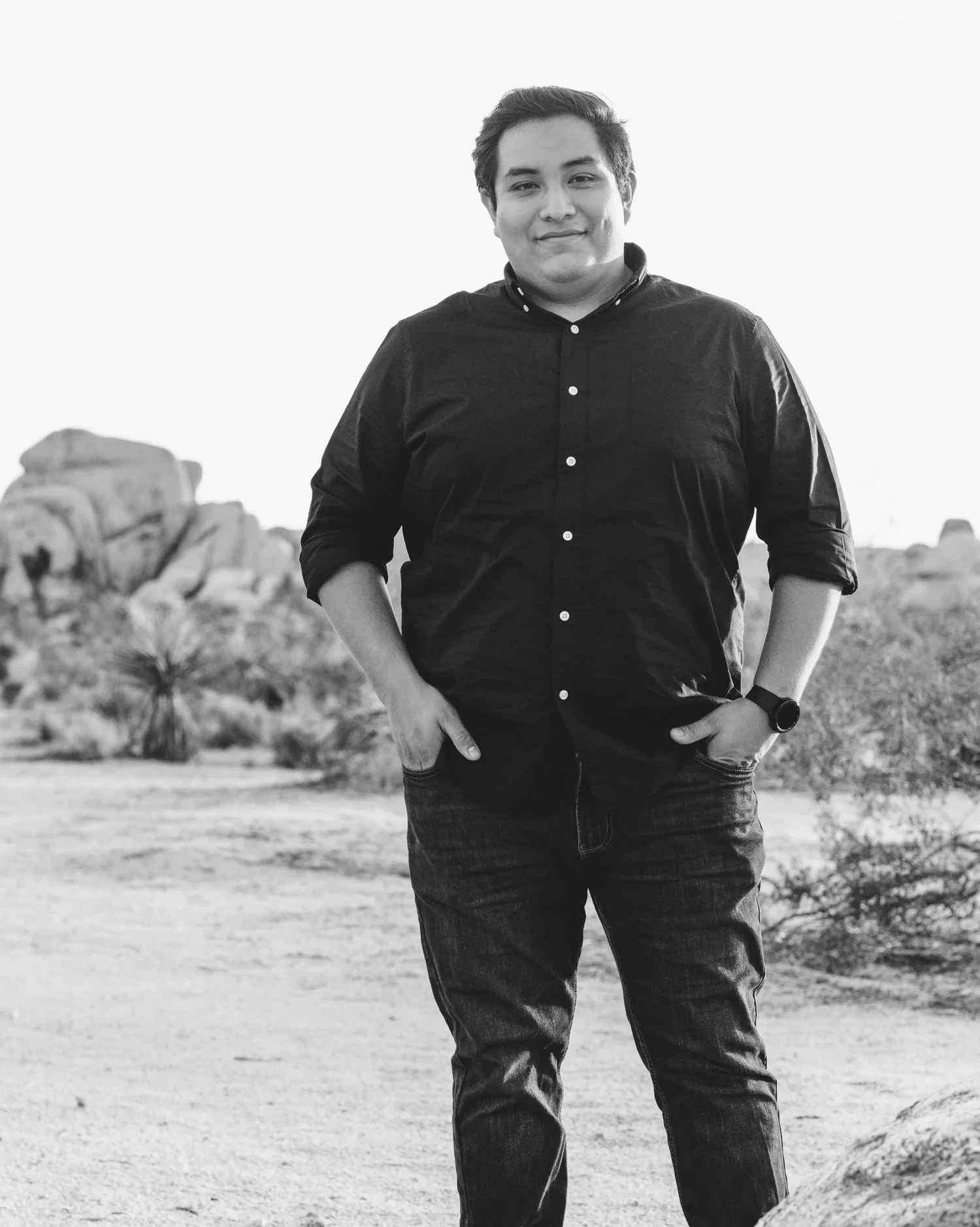 A young man with dark hair, wearing a dark button-up shirt and jeans, standing outdoors with a rocky desert landscape in the background. Black and white photo.