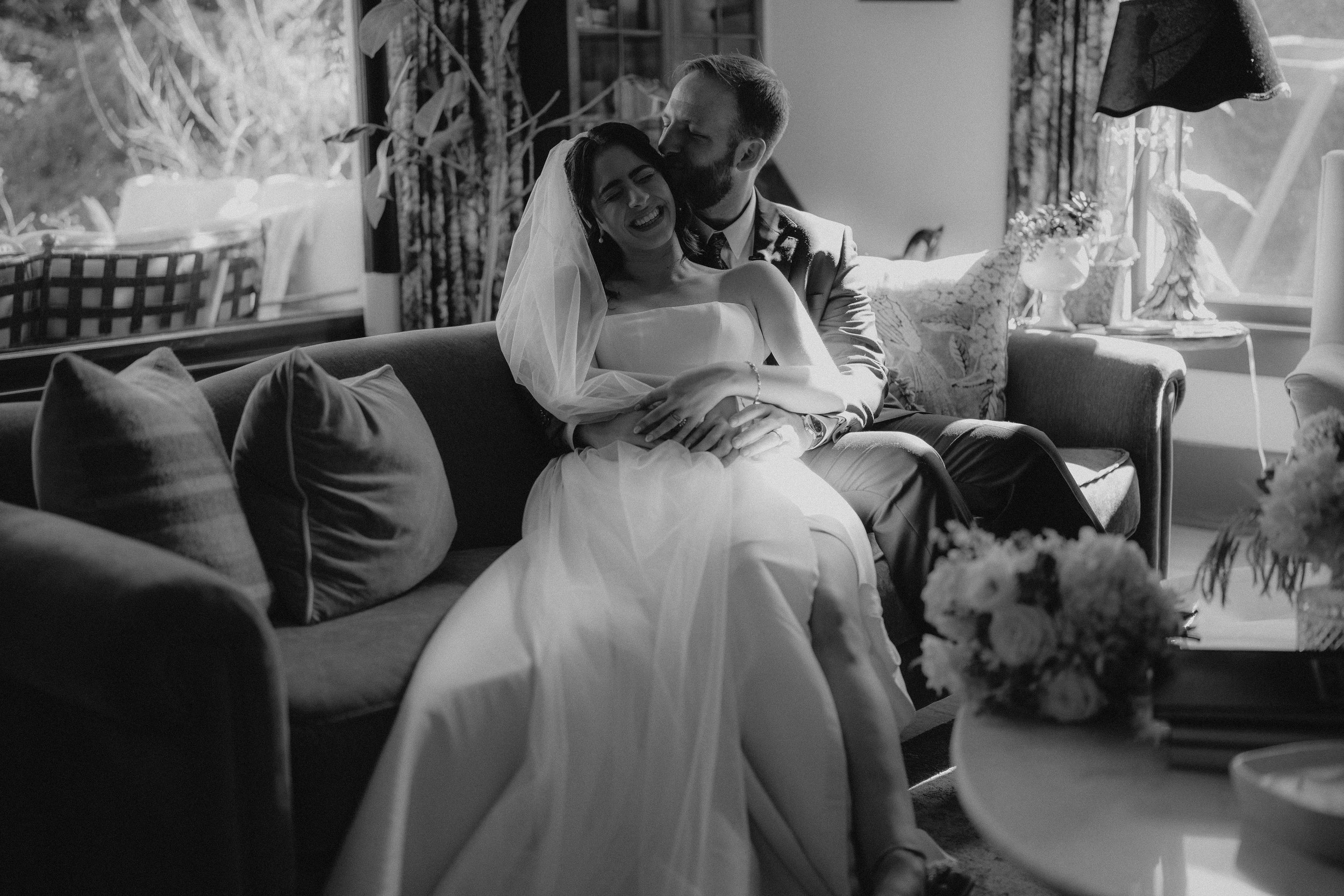 A black and white photograph of a newlywed couple sitting on a couch indoors. The bride is wearing a wedding dress and veil, and the groom is dressed in a suit. The groom is kissing the bride on her cheek, and they are smiling. There are flowers and decorative items in the background.