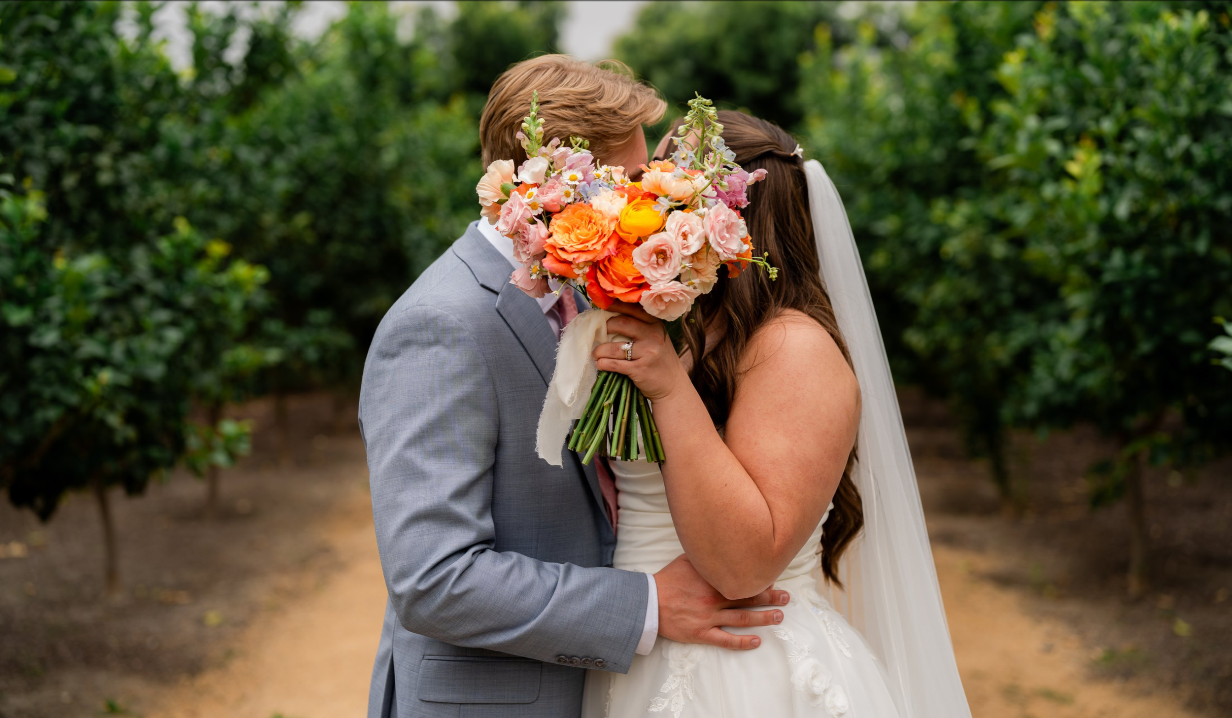 A bride and groom kissing outdoors with the bride holding a large colorful bouquet of flowers in front of their faces.