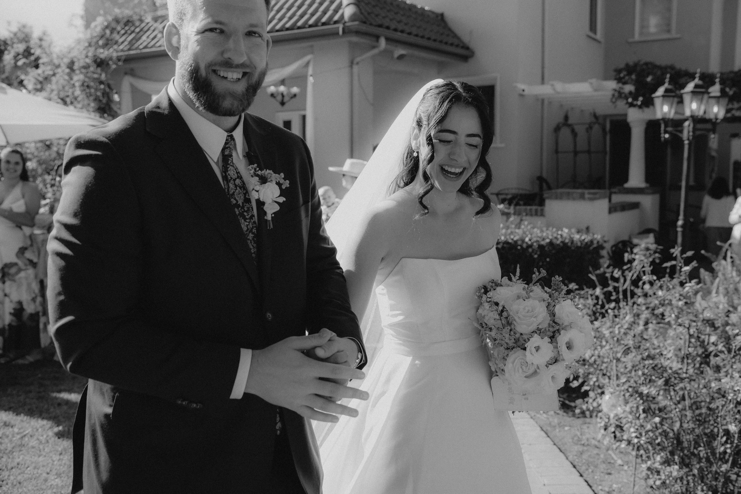 A wedding ceremony outdoors featuring a bride and groom holding hands, with the bride wearing a white wedding dress and veil, and the groom in a black suit with a floral tie. The background shows a building, trees, and guests, some of whom are seated.
