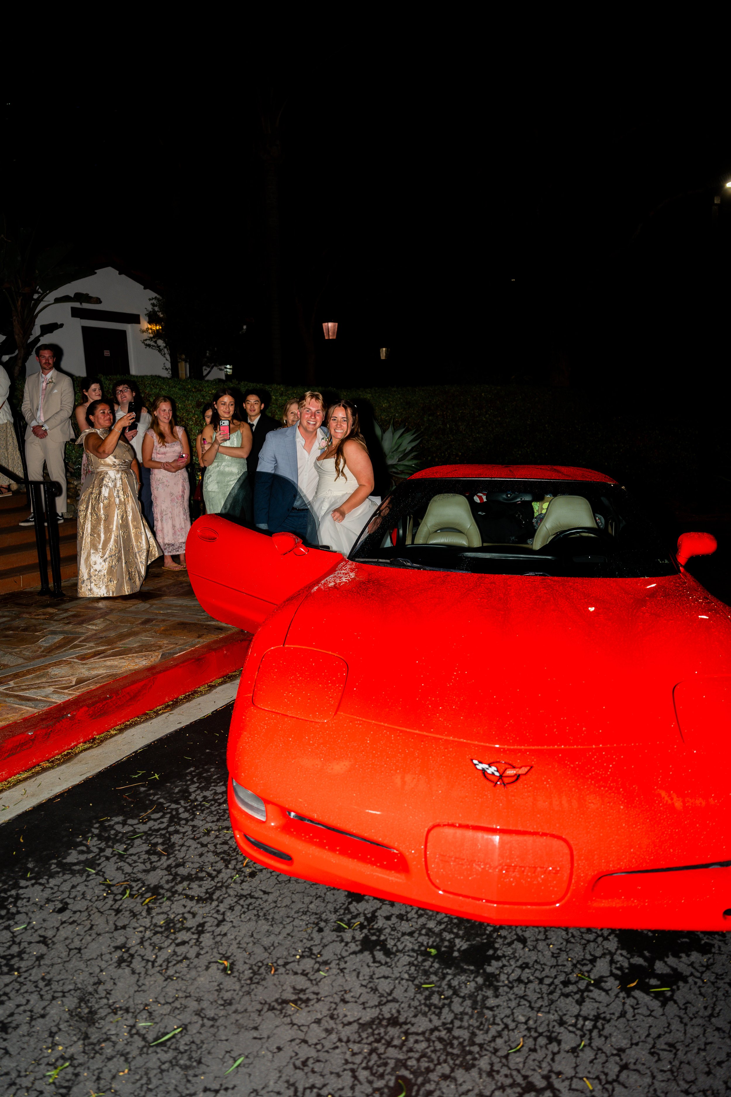 A group of people gathered outside at night, with a red sports car parked in the foreground and two people dressed in formal attire, possibly a bride and groom, smiling and posing together. The scene appears celebratory, likely from a wedding or special event.