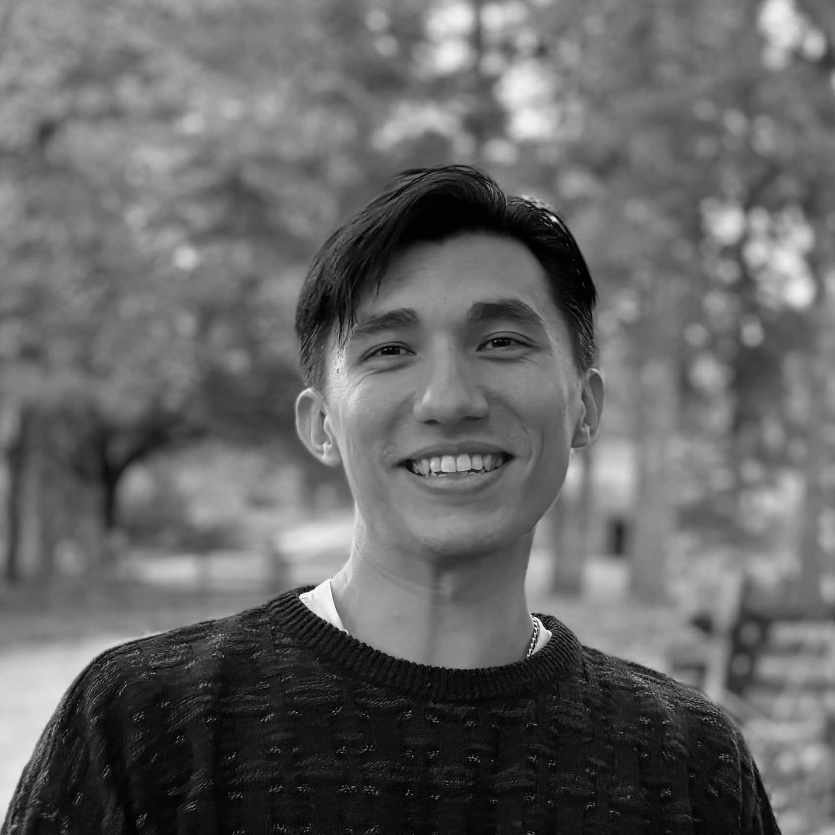 A smiling young man with dark hair standing outdoors with blurred trees in the background.