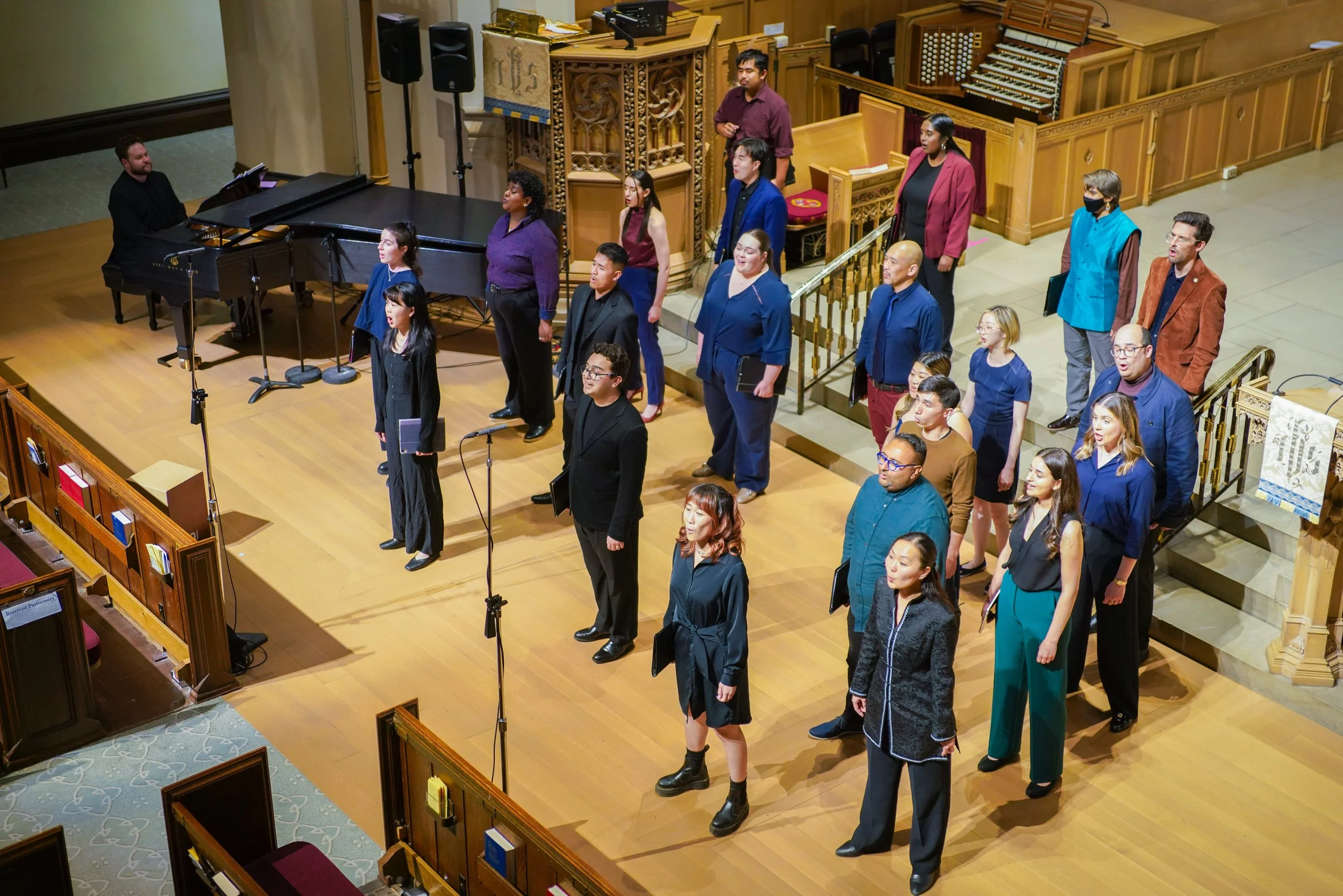 A choir standing on a wooden stage in a church, with a pianist playing a grand piano on the left, and a priest's lectern behind them. The choir members are singing, some holding sheet music, in various colorful outfits.