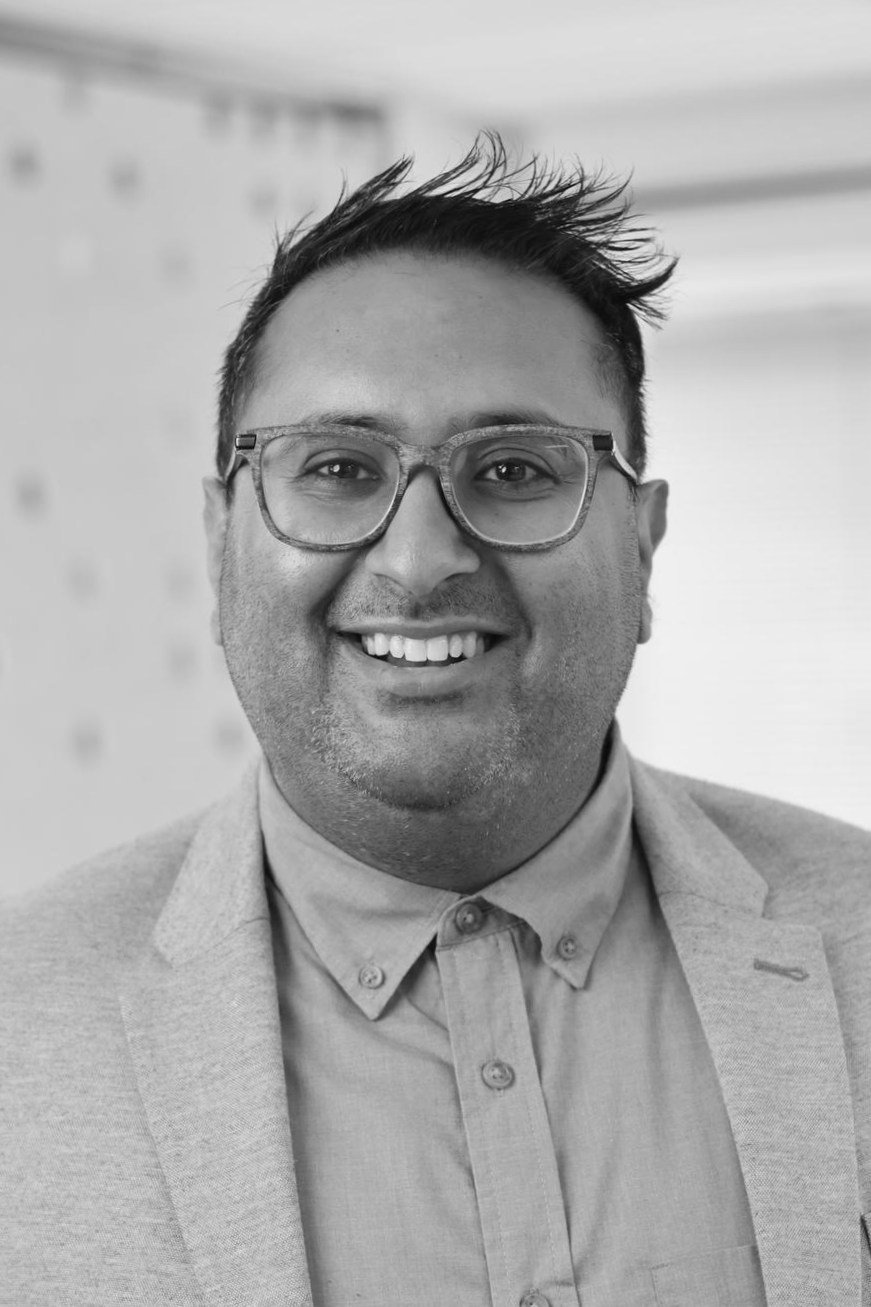 A smiling man with glasses, short hair, wearing a button-up shirt and blazer, in a black and white photo.
