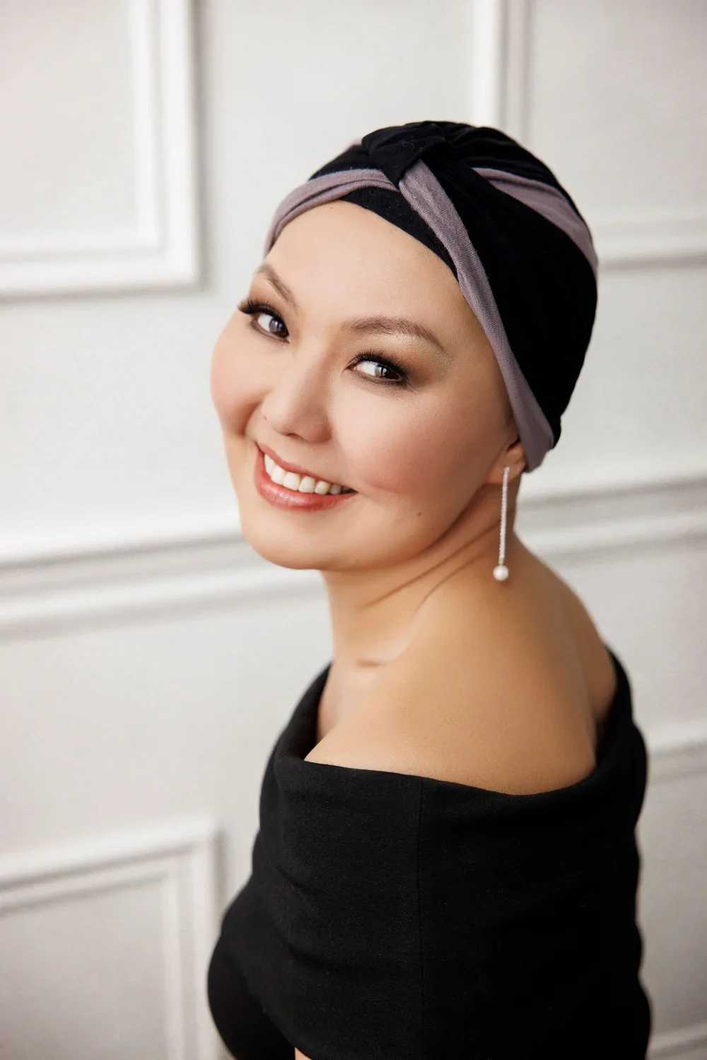 A woman with light skin, smiling, wearing a black off-the-shoulder top, a grey and black headscarf, and a dangling pearl earring, standing indoors against a white wall.