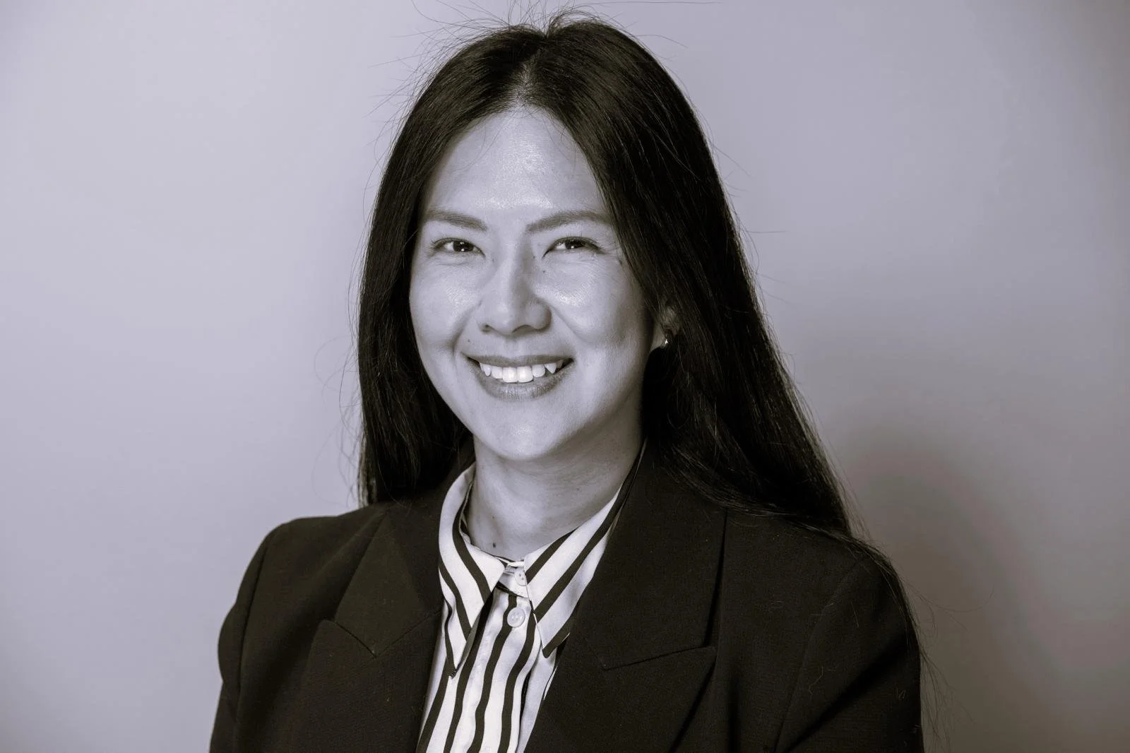 Black and white portrait of a smiling woman with long dark hair, wearing a black blazer and a striped blouse, against a plain background.
