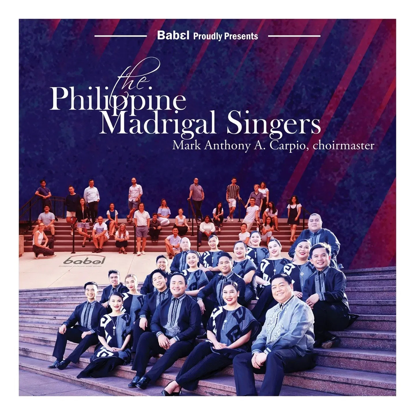 Group photo of Philippine Madrigal Singers with members in traditional Filipino attire seated on stairs, with others standing behind on an outdoor stage at night.