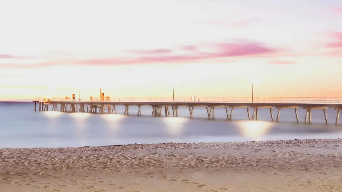 Long pier extending into the ocean at sunset, with a sandy beach in the foreground and pink-tinged clouds in the sky.
