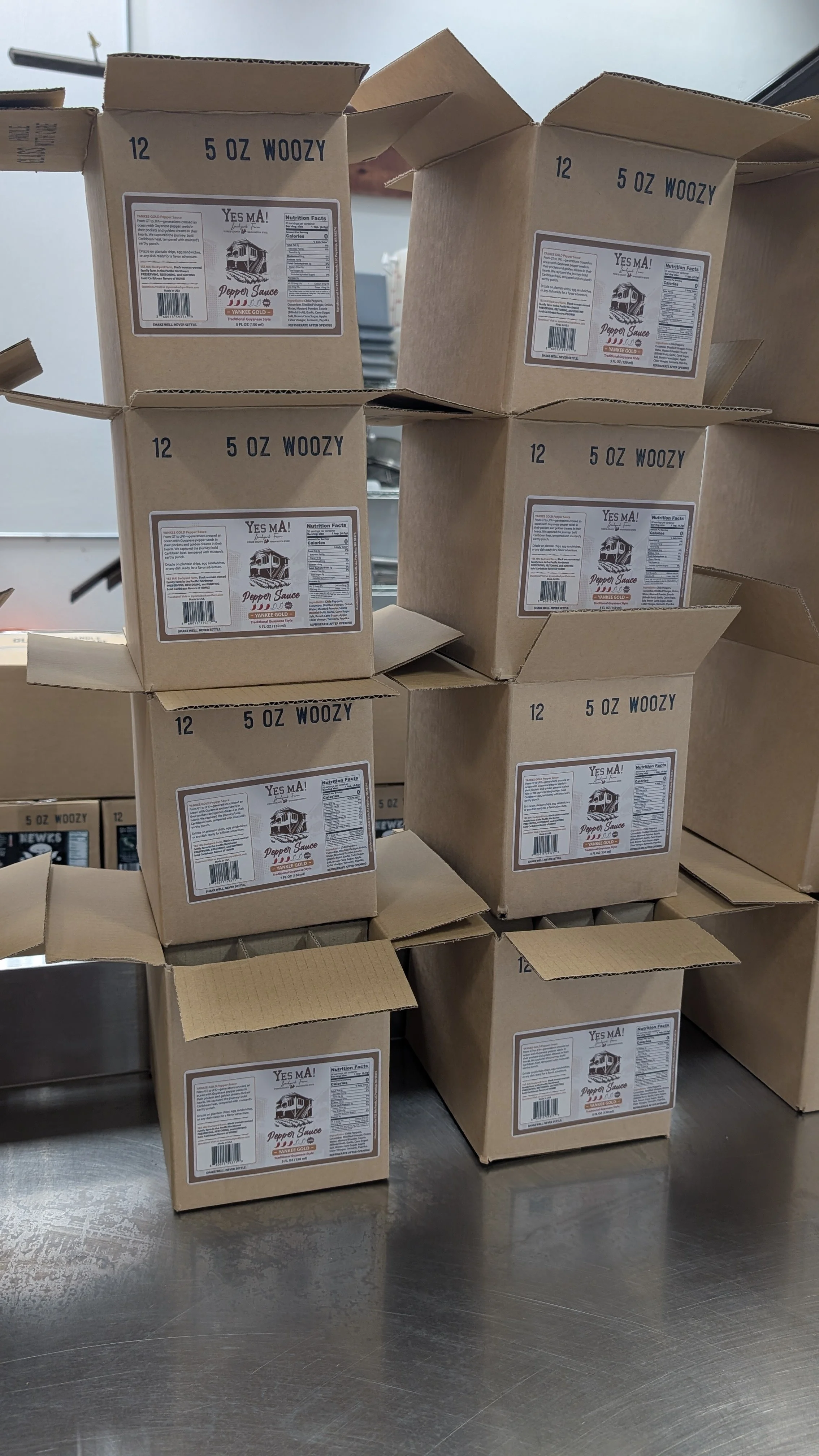 Stacked open cardboard boxes labeled '12 5 OZ WOOZY' with a 'Pepper Sauce' label on them, on a metal surface in a warehouse or storage area.