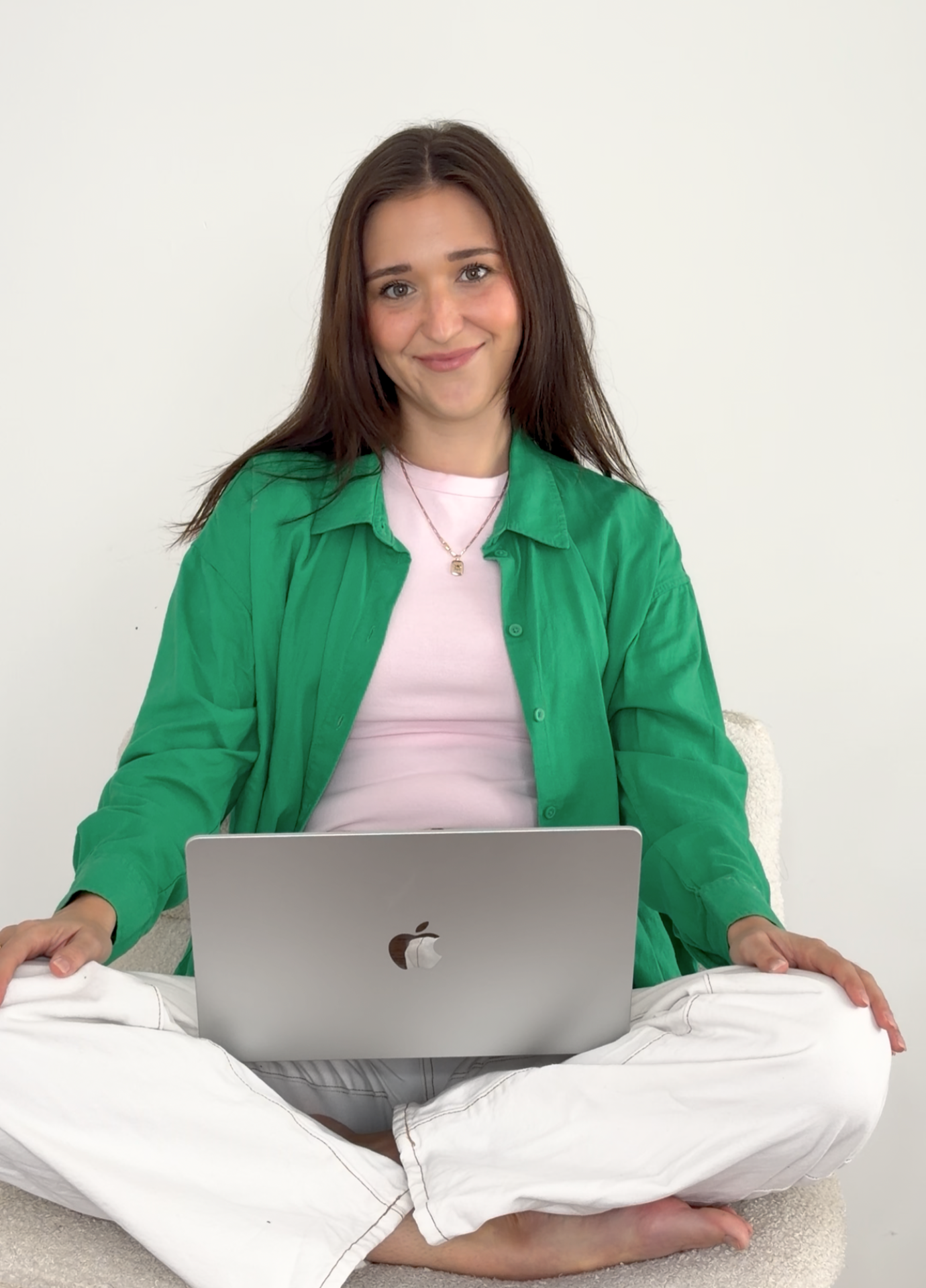 A young woman with long brown hair sitting cross-legged on the floor, smiling, using a silver MacBook laptop. She is wearing a green shirt over a white t-shirt, white pants, and a necklace.