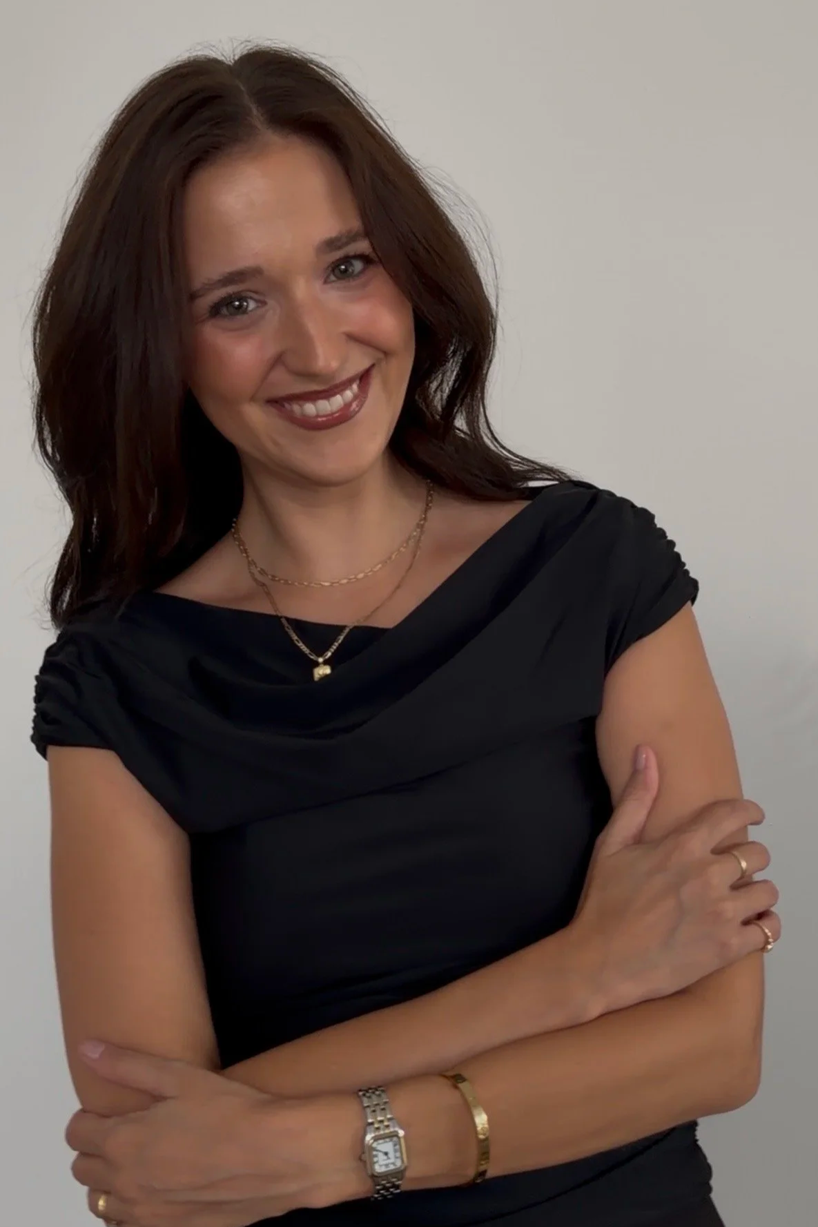 A woman with brown hair wearing a black top, gold jewelry, and a watch, smiling with her arms crossed against a plain white background.