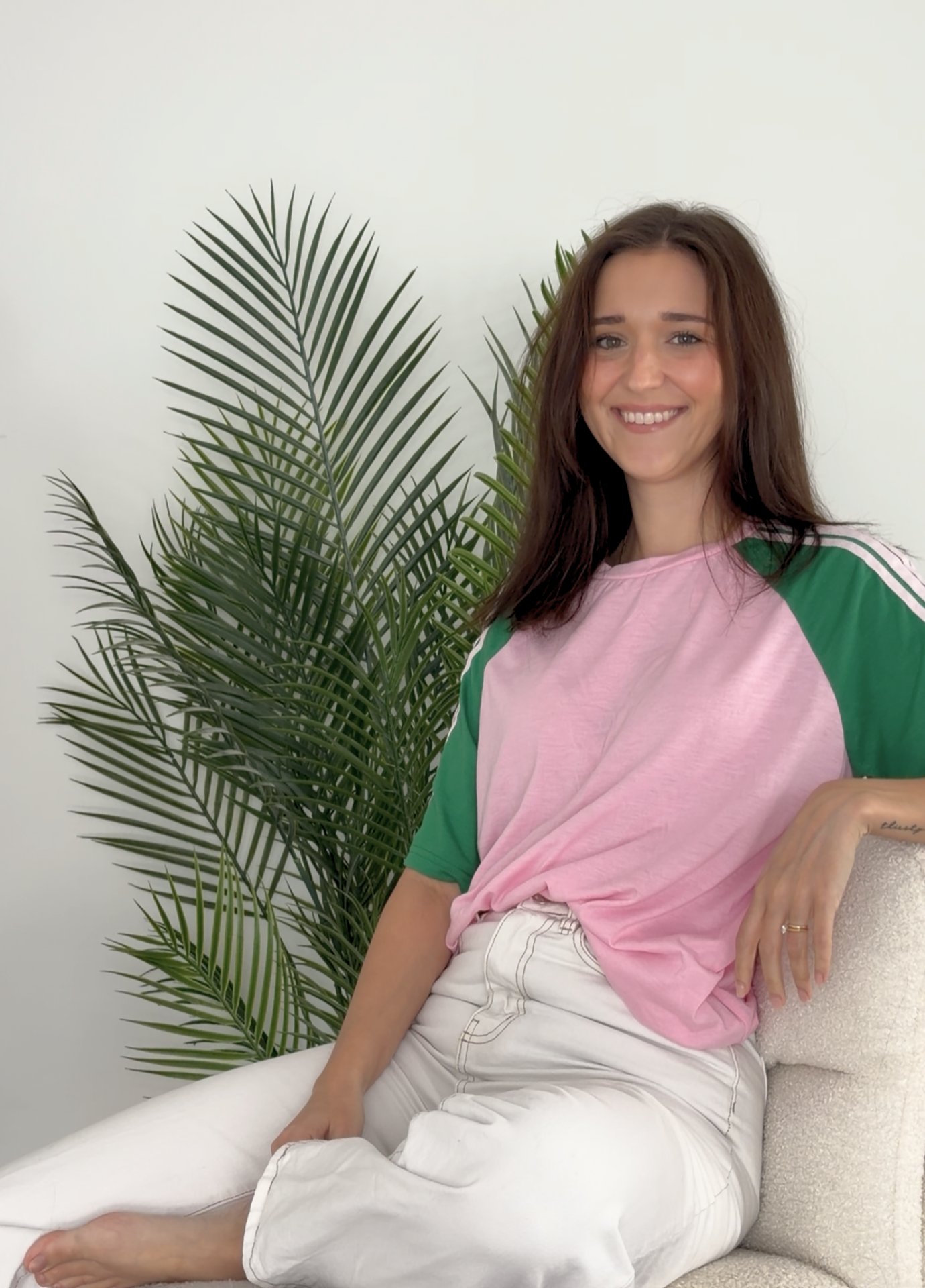 A young woman with brown hair and a pink and green t-shirt sitting on a cream-colored sofa, smiling, with large green potted plants in the background against a plain light-colored wall.