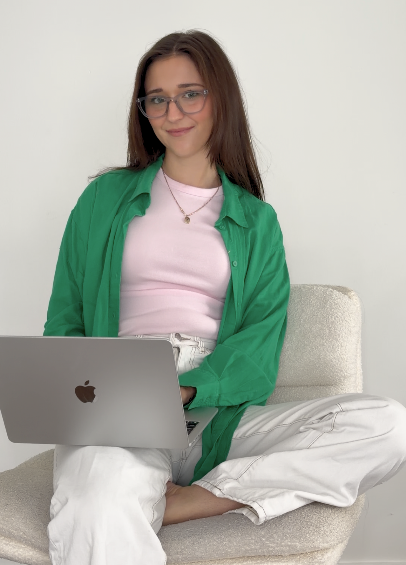A young woman with long brown hair, glasses, and a friendly expression, sitting cross-legged on a beige textured chair with a silver MacBook on her lap, wearing a pink T-shirt, green unbuttoned shirt, and white pants against a plain white wall background.