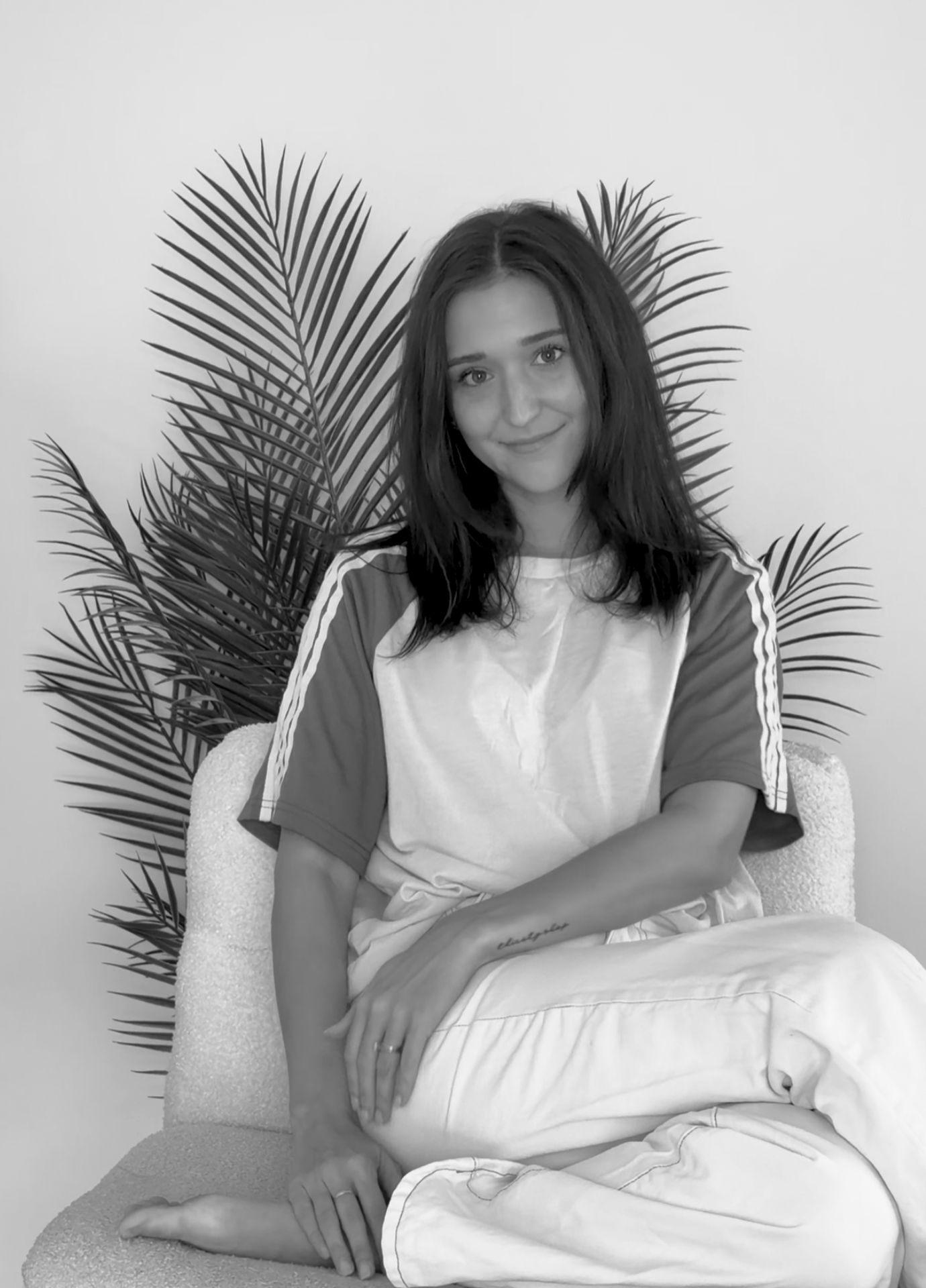 A young woman with dark hair sitting on a chair in front of large palm leaves, smiling at the camera. The photo is in black and white.