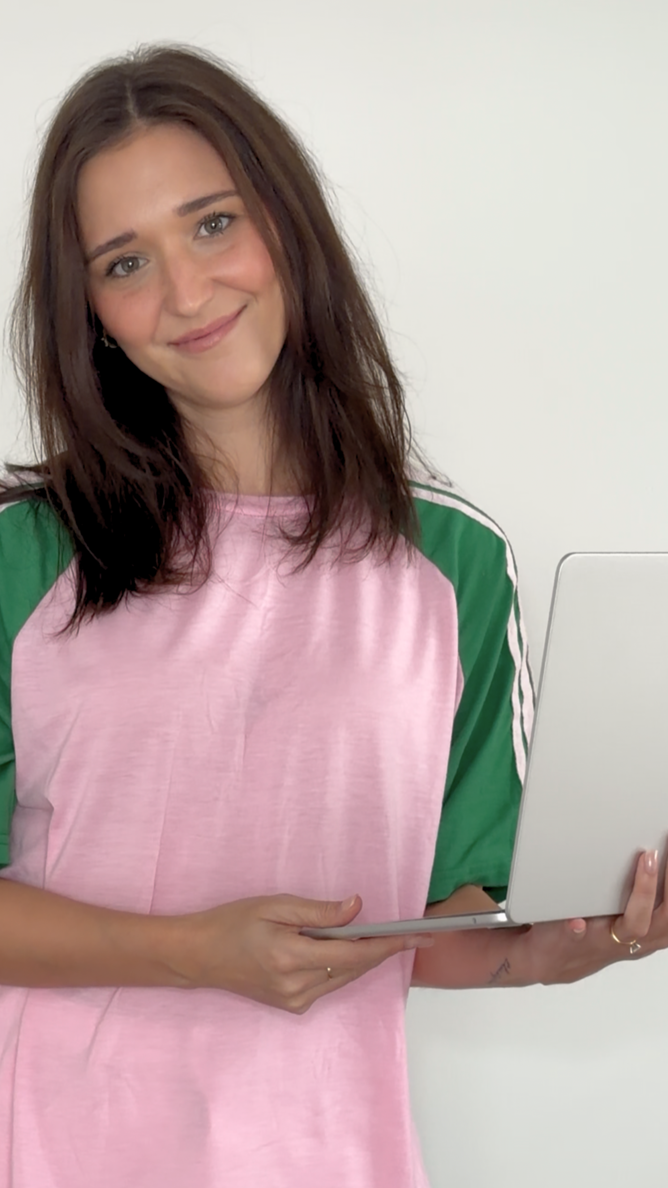 A young woman with long brown hair, wearing a pink and green shirt, holding a silver laptop, smiling, standing against a plain white wall.