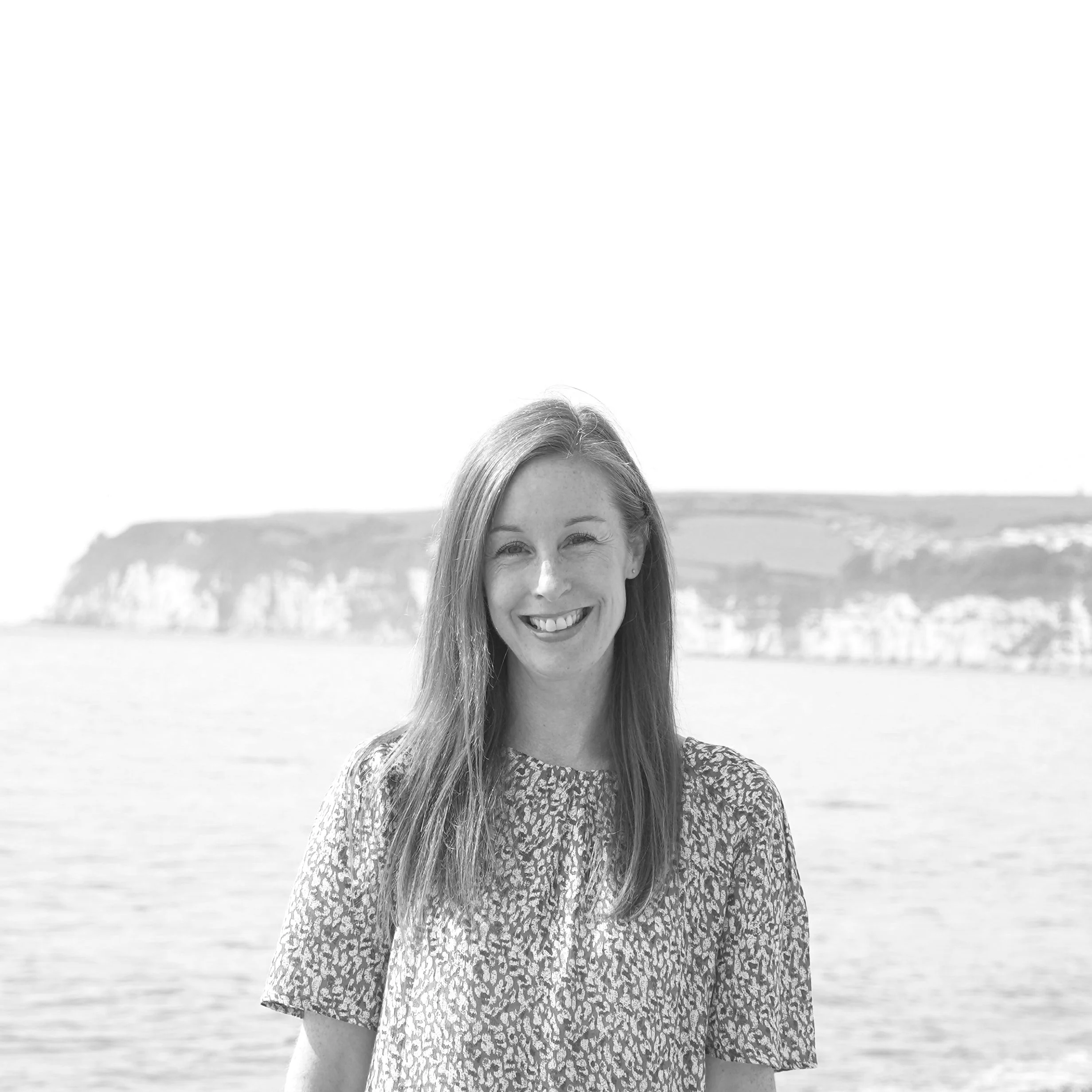 A woman with long hair smiling outdoors near water, with cliffs in the background.